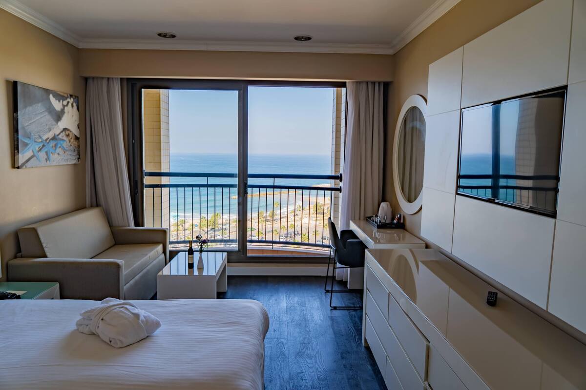 Habitación de hotel con cama, sofá, escritorio, televisor y balcón con vista al mar.