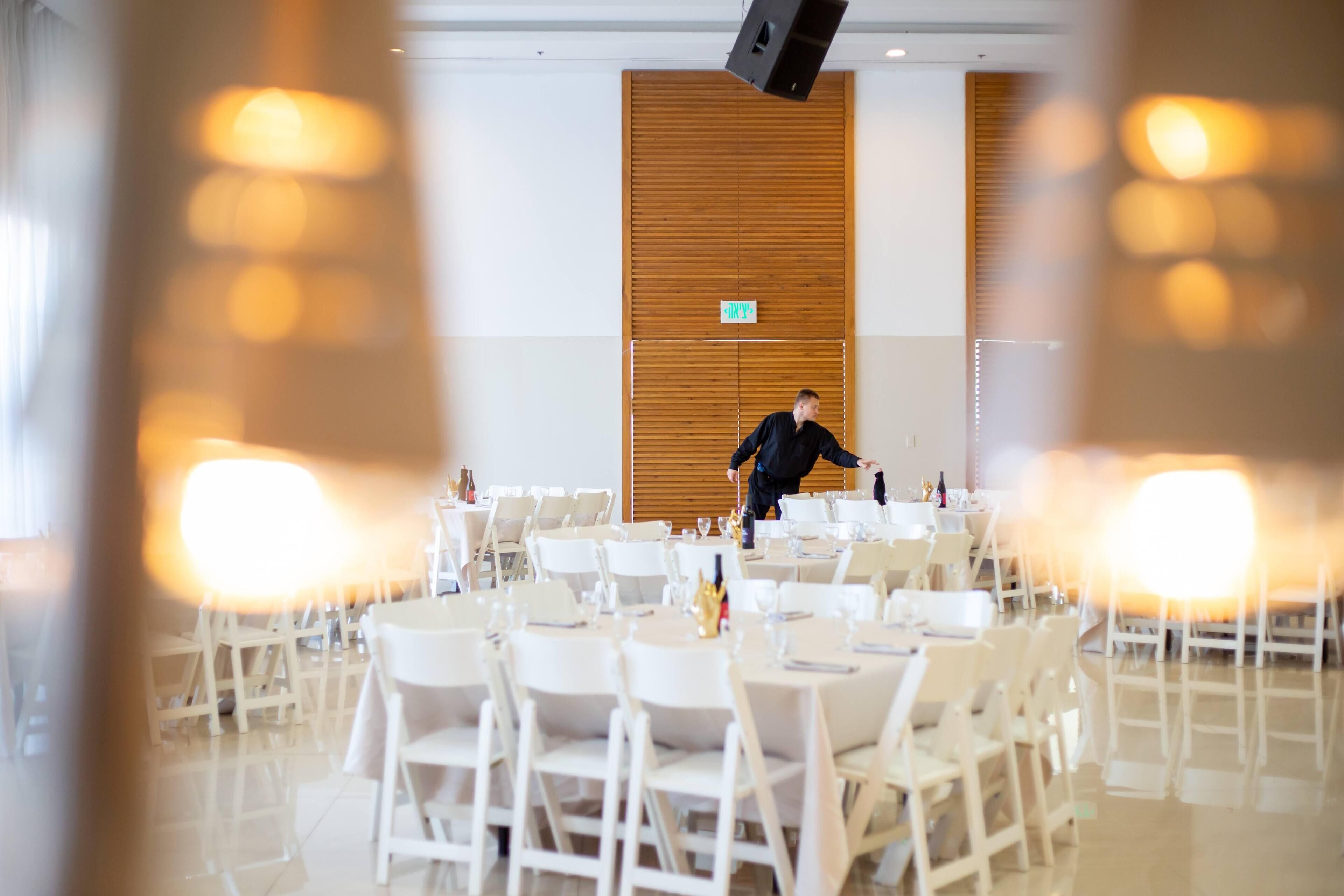Un amplio salón de banquetes con mesas y sillas blancas, un panel de pared de madera y un letrero que indica una salida.