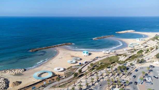 An aerial view of a coastal area with a beach, a rocky breakwater, and a parking lot with cars. - Tamara Ashkelon (Ashkelon)