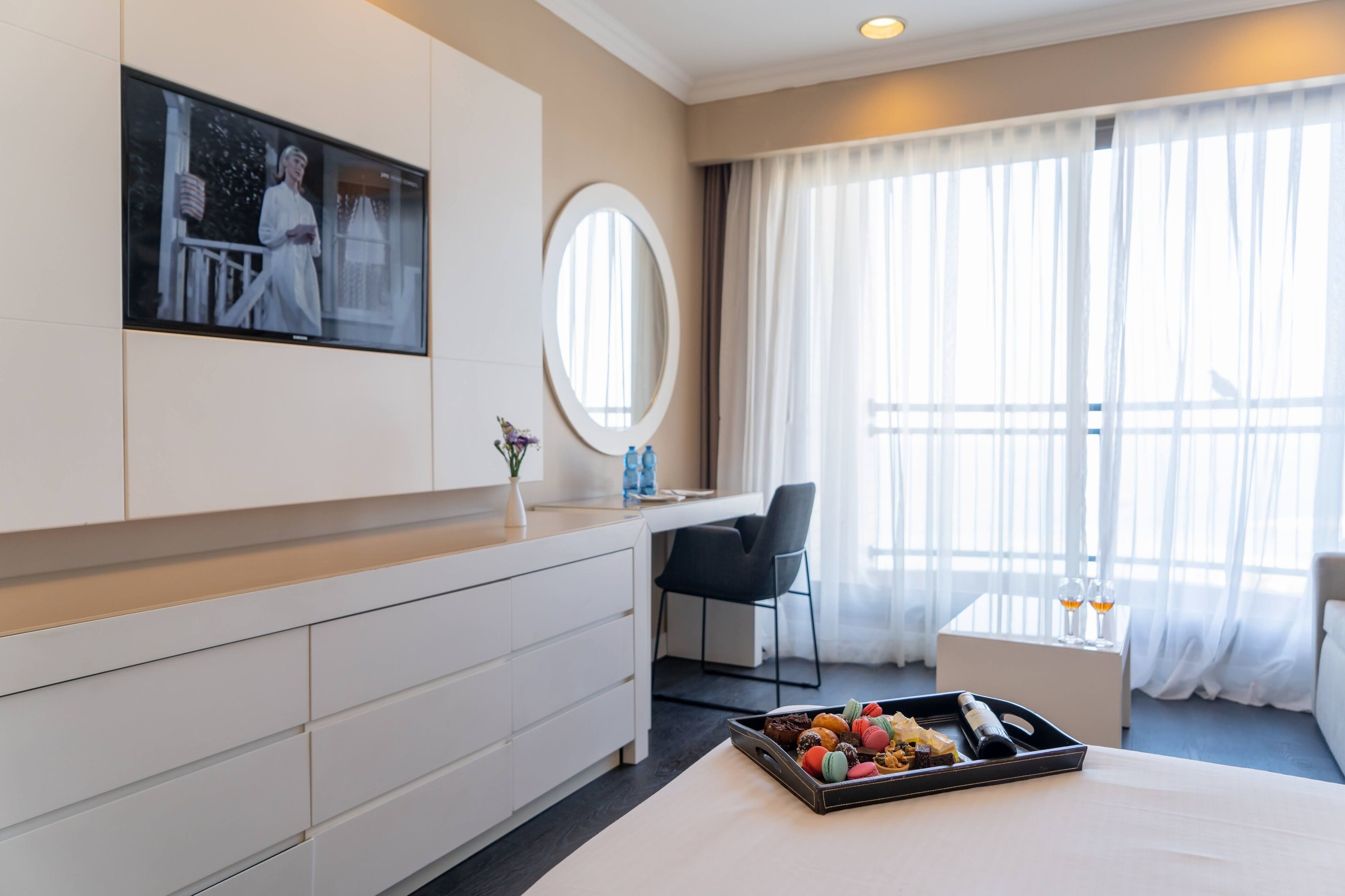 A hotel room with a bed, a desk, a chair, a TV, a mirror, a window with curtains, and a tray with snacks.