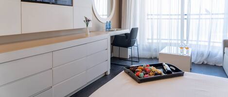 A hotel room with a bed, a desk, a chair, a TV, a mirror, a window with curtains, and a tray with snacks.
