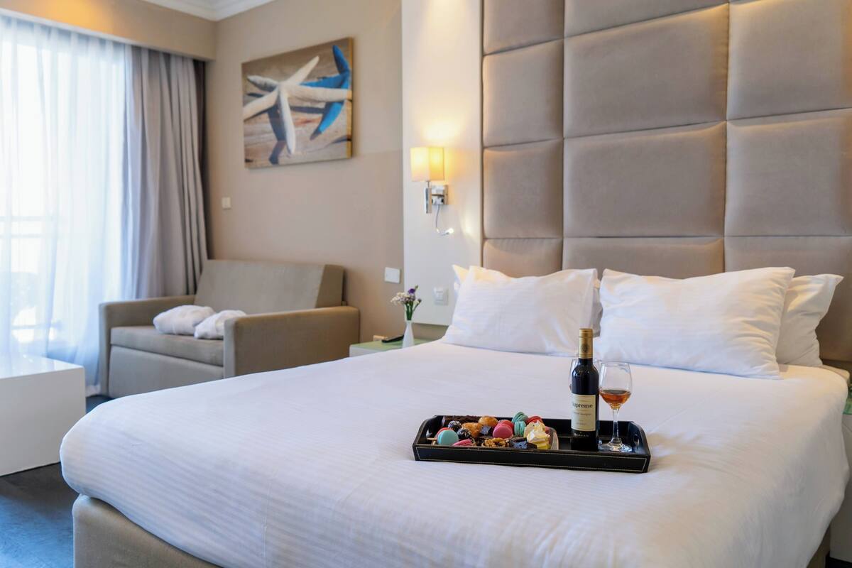 Habitación de hotel con una cama grande, una silla, una mesita de noche y una bandeja con una botella y vasos.
