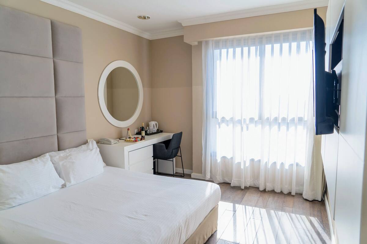 Habitación de hotel con una cama grande, un escritorio con silla, un espejo redondo y un televisor montado en la pared.