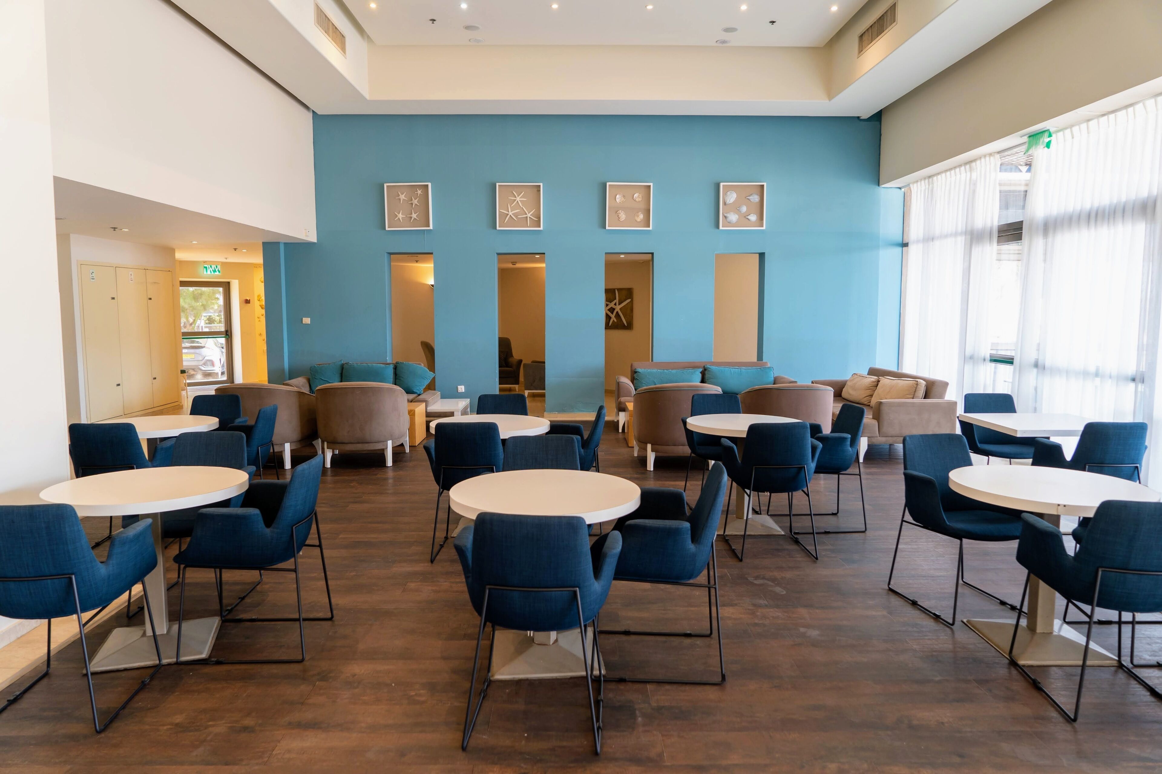 A spacious indoor area with multiple round tables and blue chairs, featuring a blue wall with framed artwork and a large window with sheer curtains.