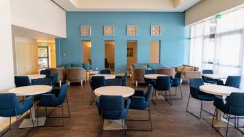 A spacious indoor area with multiple round tables and blue chairs, featuring a blue wall with framed artwork and a large window with sheer curtains.
