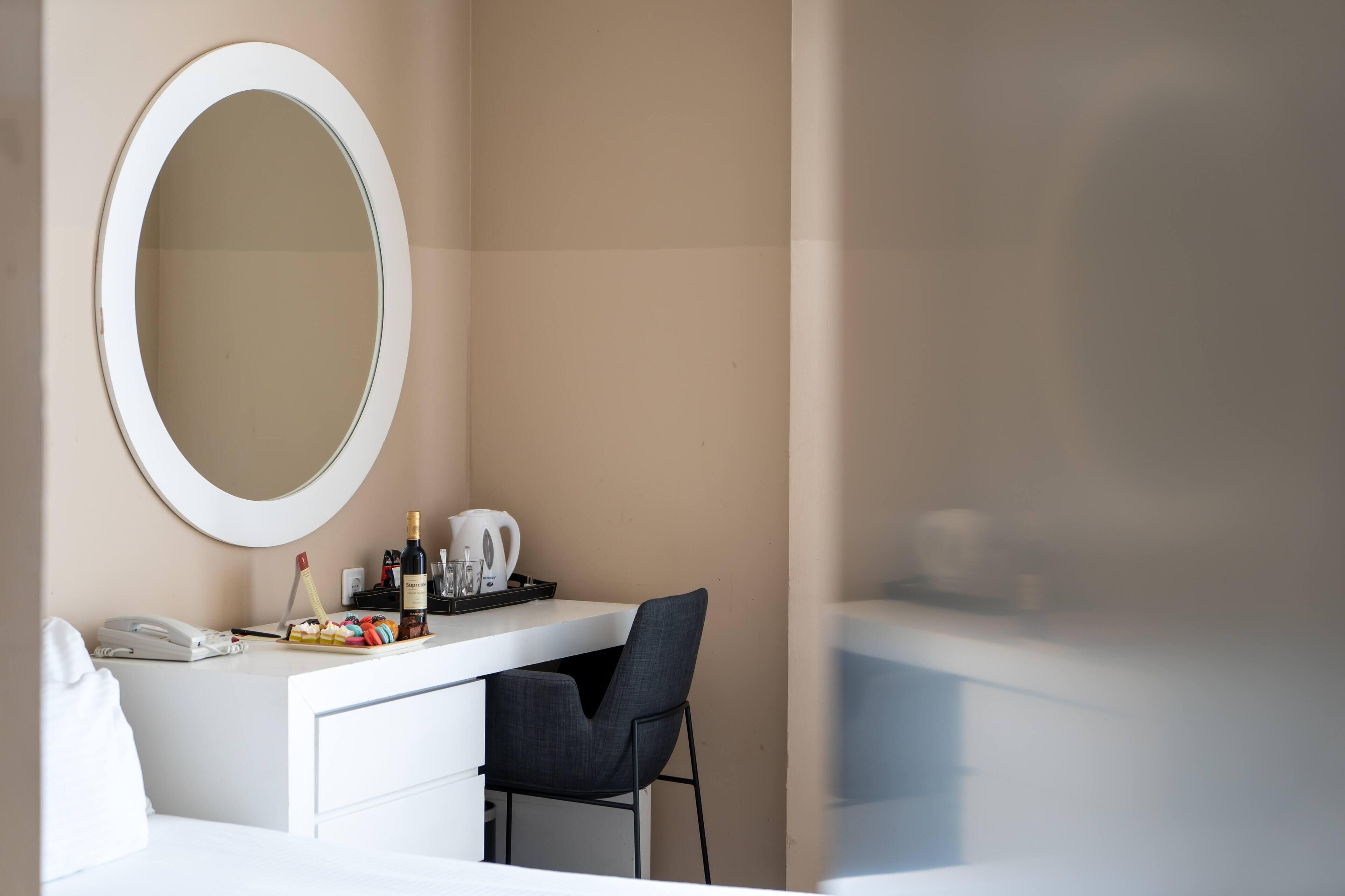 A modern hotel room with a white vanity, a round mirror, a desk with a chair, and a minibar.
