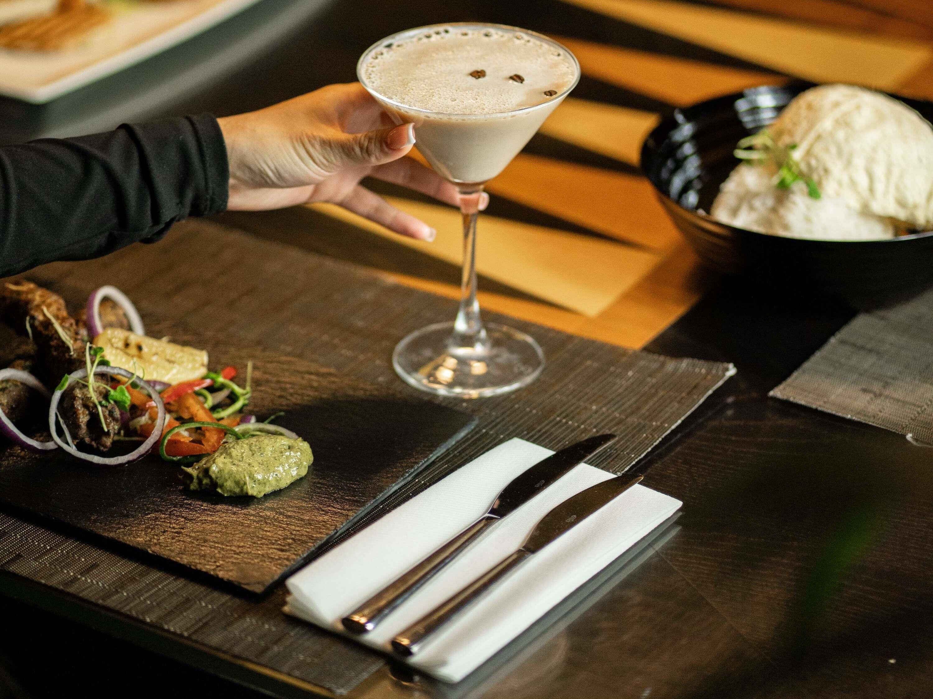 A person holding a martini glass with a frothy drink, a plate of food including grilled meat and vegetables, and a bowl of soup on a wooden table.