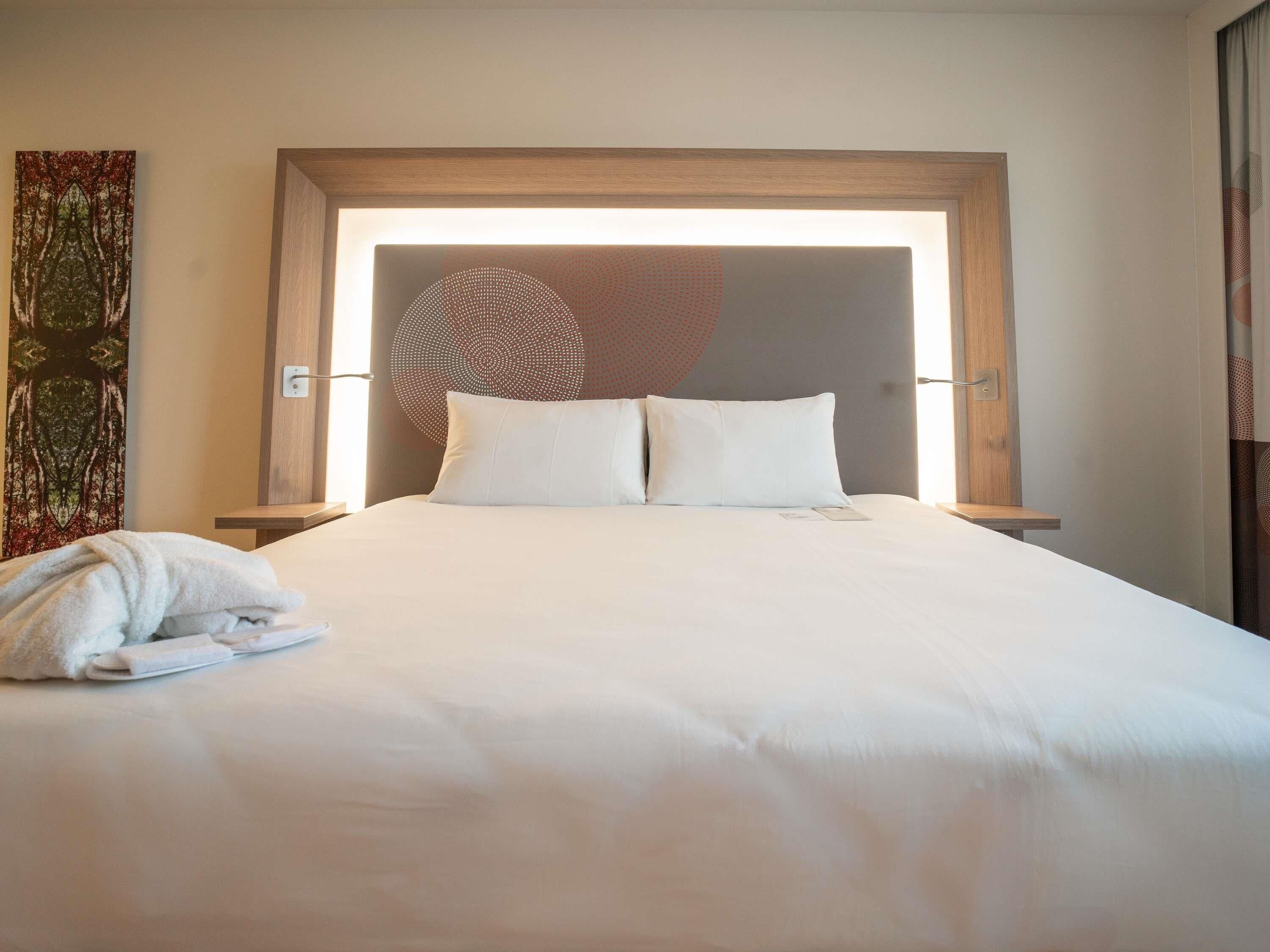 Un lit bien fait, avec du linge de lit blanc, une tête de lit en bois et un luminaire fixé au mur.