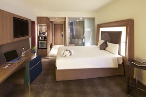 Habitación de hotel con cama, escritorio con computadora portátil, televisión, refrigerador pequeño y zona de ducha.