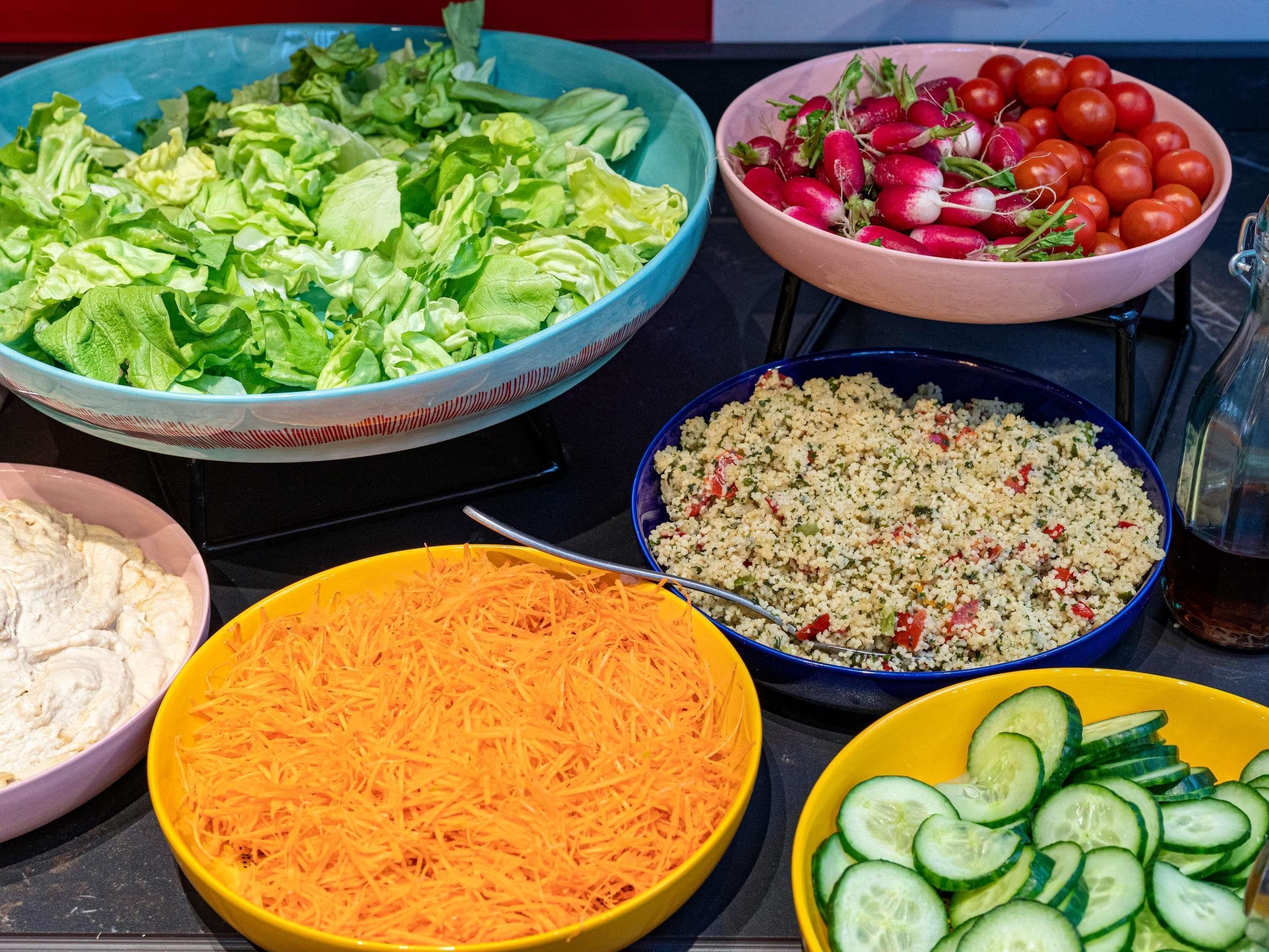A table with various food items including a bowl of shredded carrots, a bowl of sliced cucumbers, a bowl of couscous salad, a bowl of lettuce, a bowl of radishes, and a bowl of tomatoes.