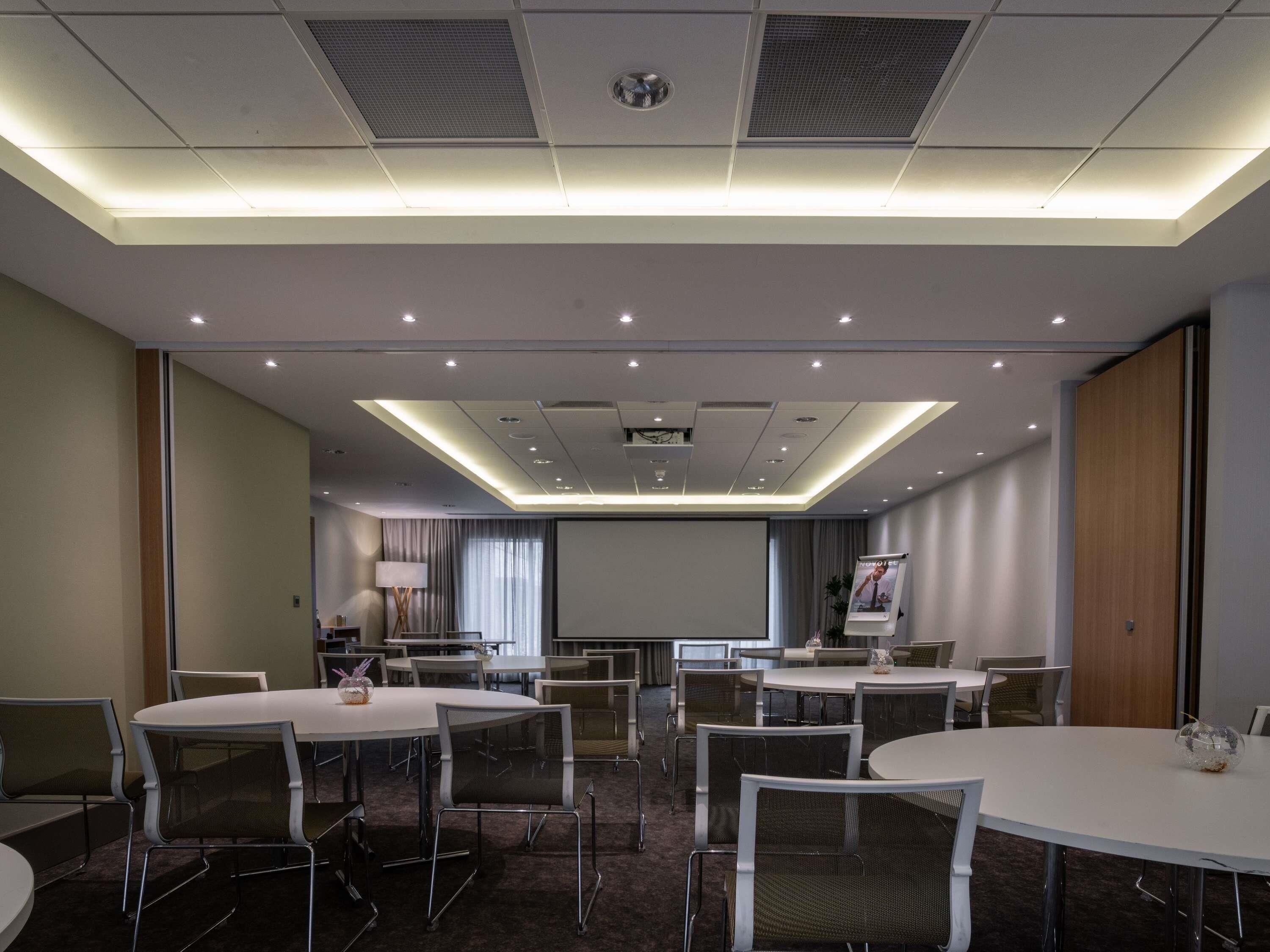 A conference room with multiple tables and chairs, a projector screen, and a ceiling with recessed lighting.