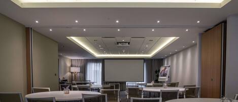 Un salón de conferencias con varias mesas y sillas, una pantalla de proyector e iluminación empotrada en el techo.