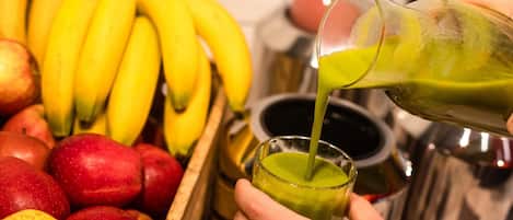 Una mano vertiendo un líquido verde en un vaso, con un cajón de madera con frutas y un recipiente metálico al fondo.