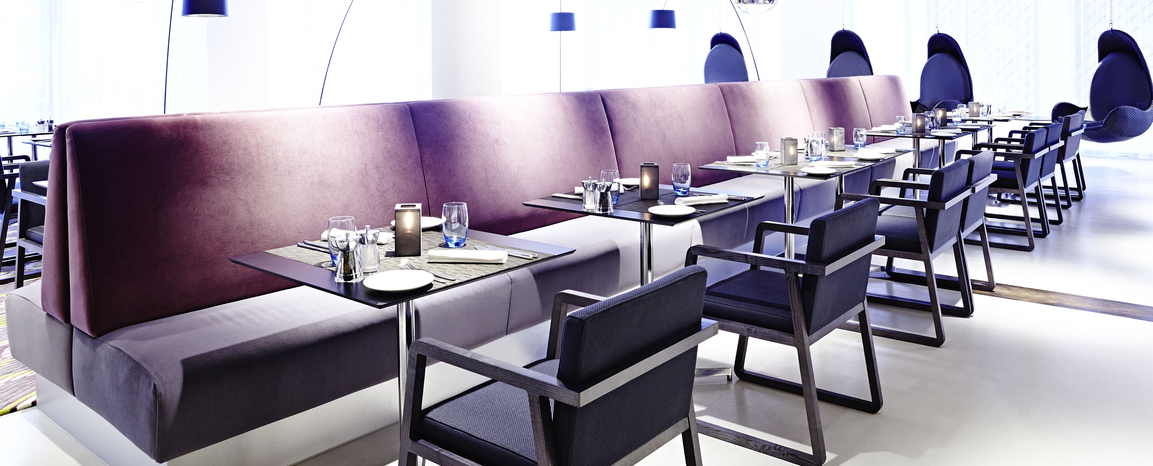 A modern restaurant with purple booths and black chairs, featuring neatly arranged tables with glassware and cutlery.