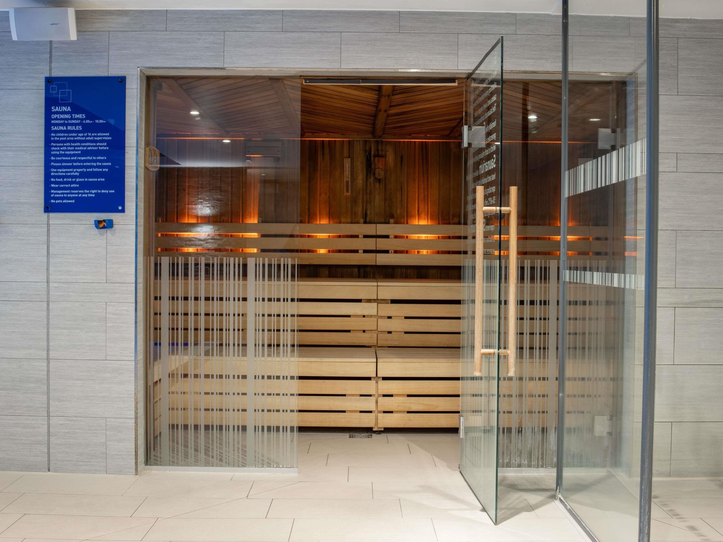 A modern sauna with a glass entrance, wooden benches, and a sign detailing sauna rules and opening times.