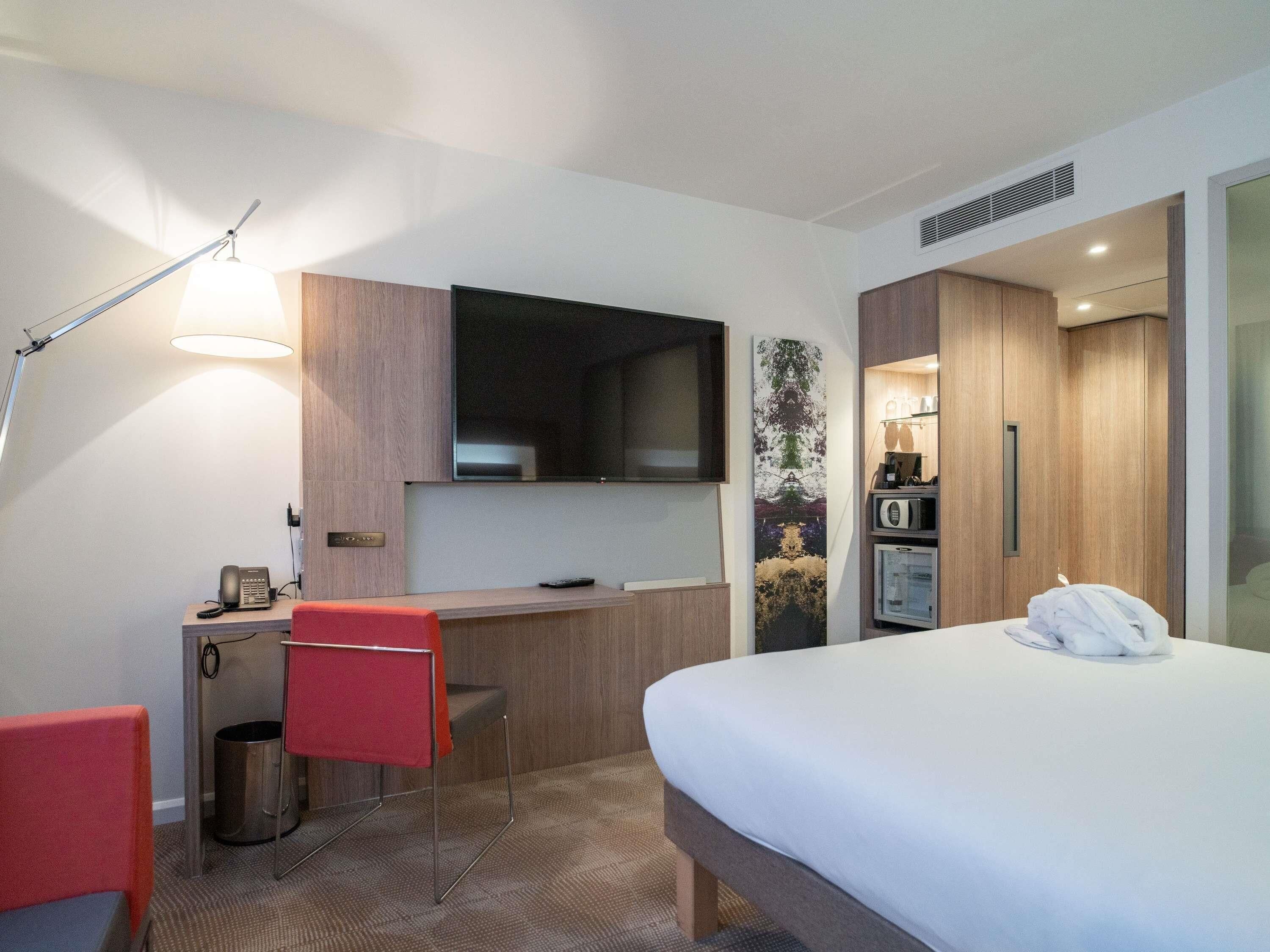 Habitación de hotel con cama, escritorio con silla, televisor montado en la pared, microondas y un cubo de basura pequeño.