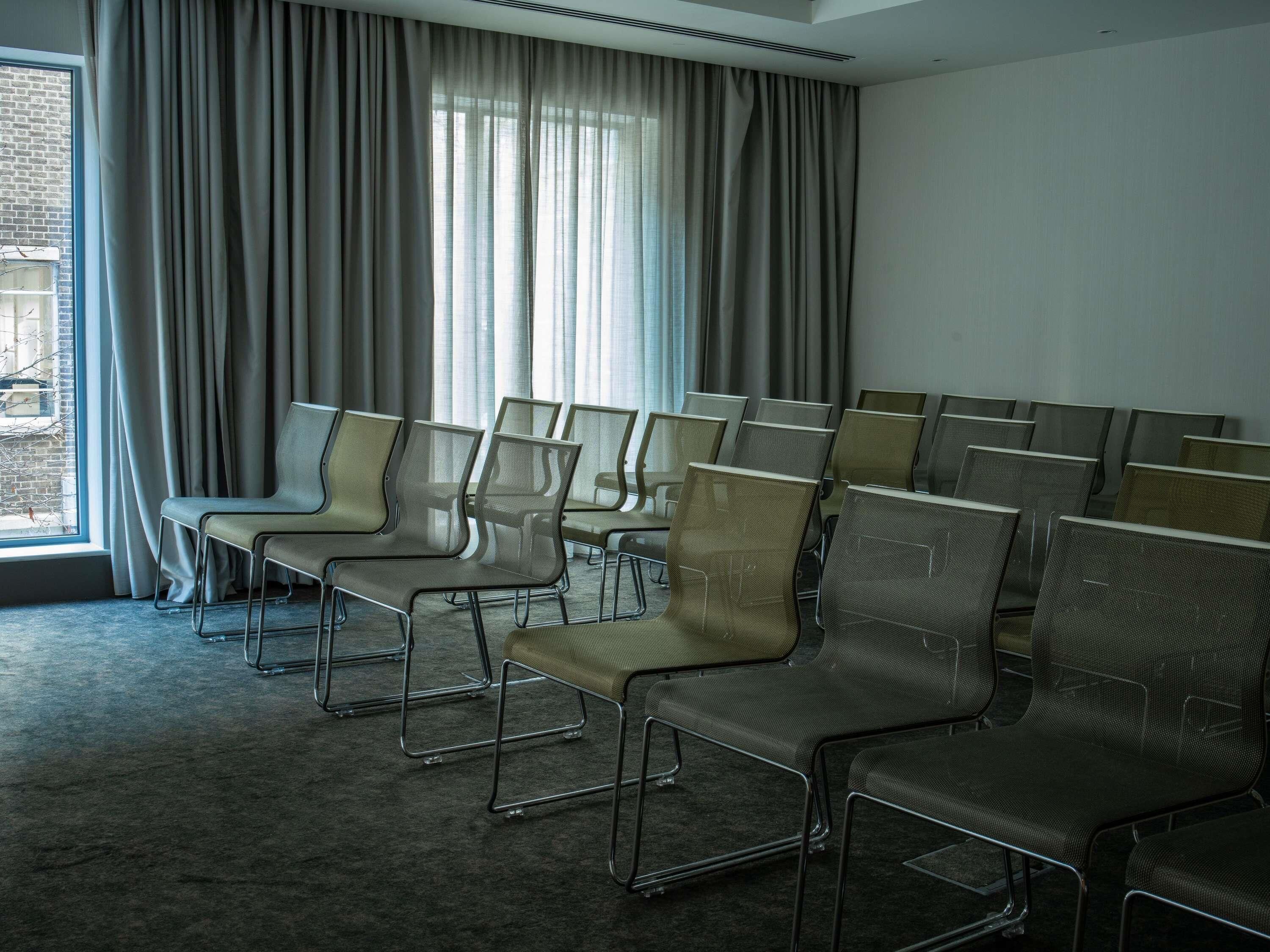 Ein Konferenzraum mit Reihen von Stühlen, großen Fenstern mit Vorhängen und Blick auf ein Gebäude.