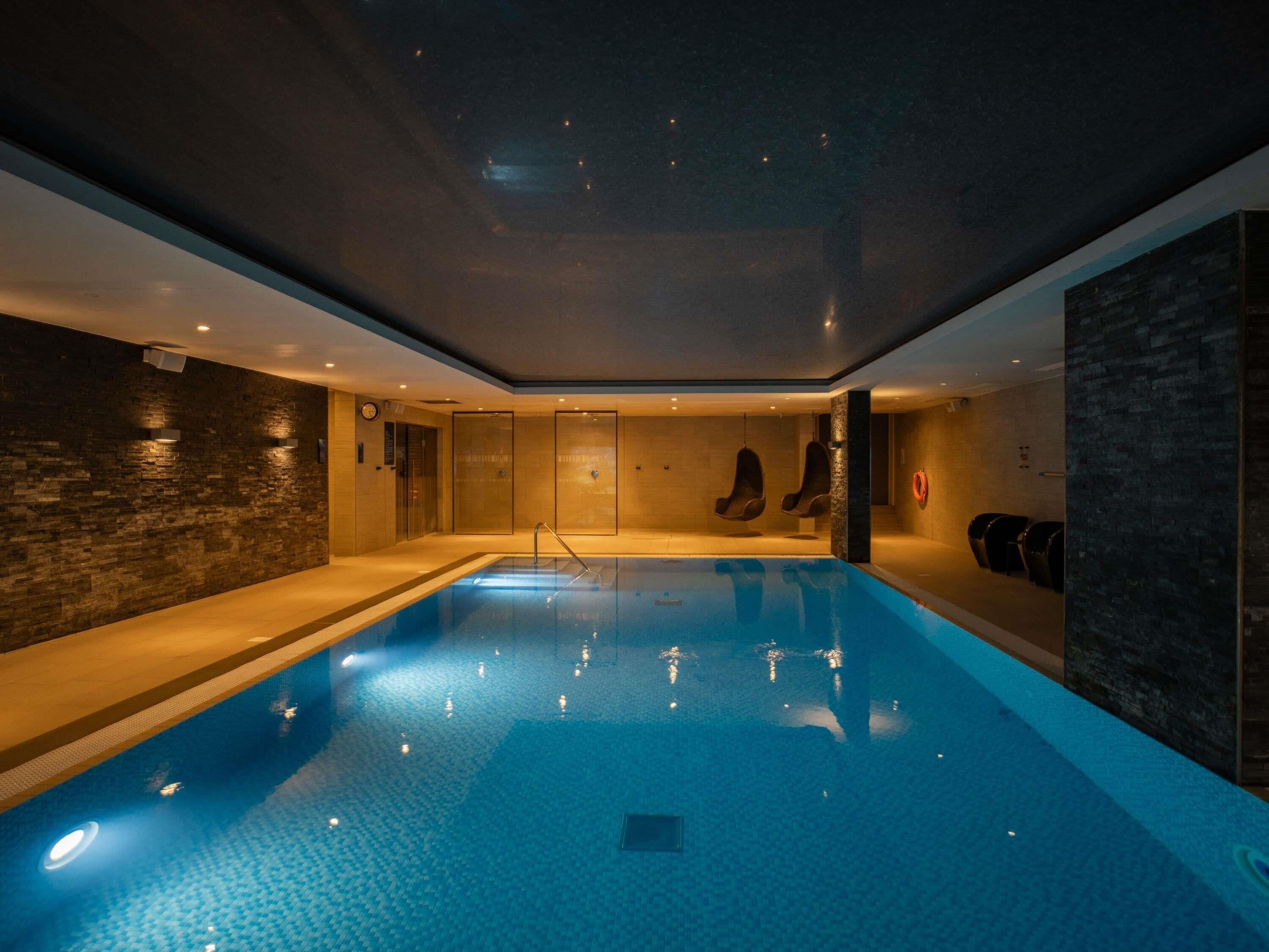A swimming pool with a tiled floor, a dark wall with a clock, and a hanging chair.