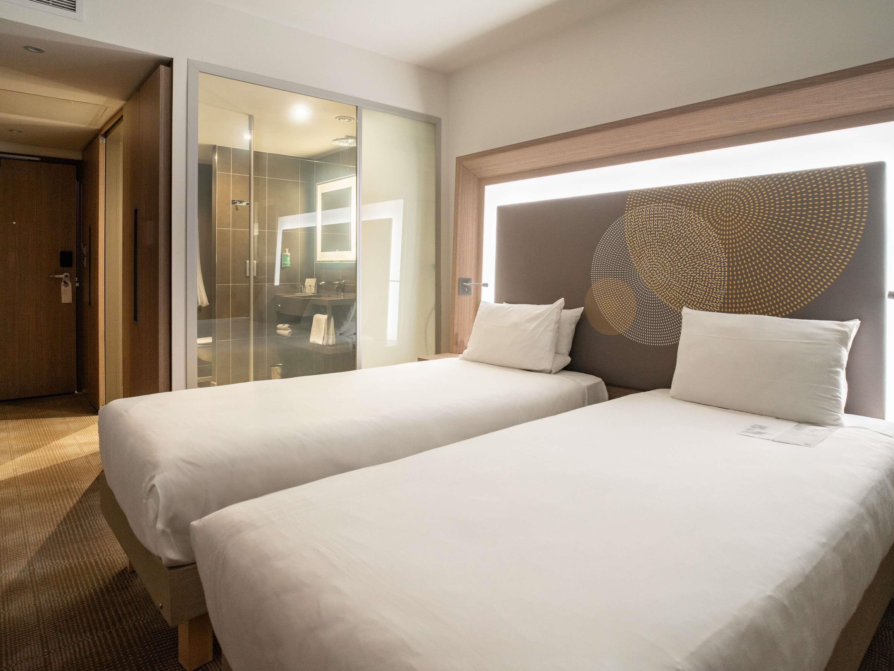 A hotel room with two beds, a glass shower enclosure, and a modern headboard with a circular design.