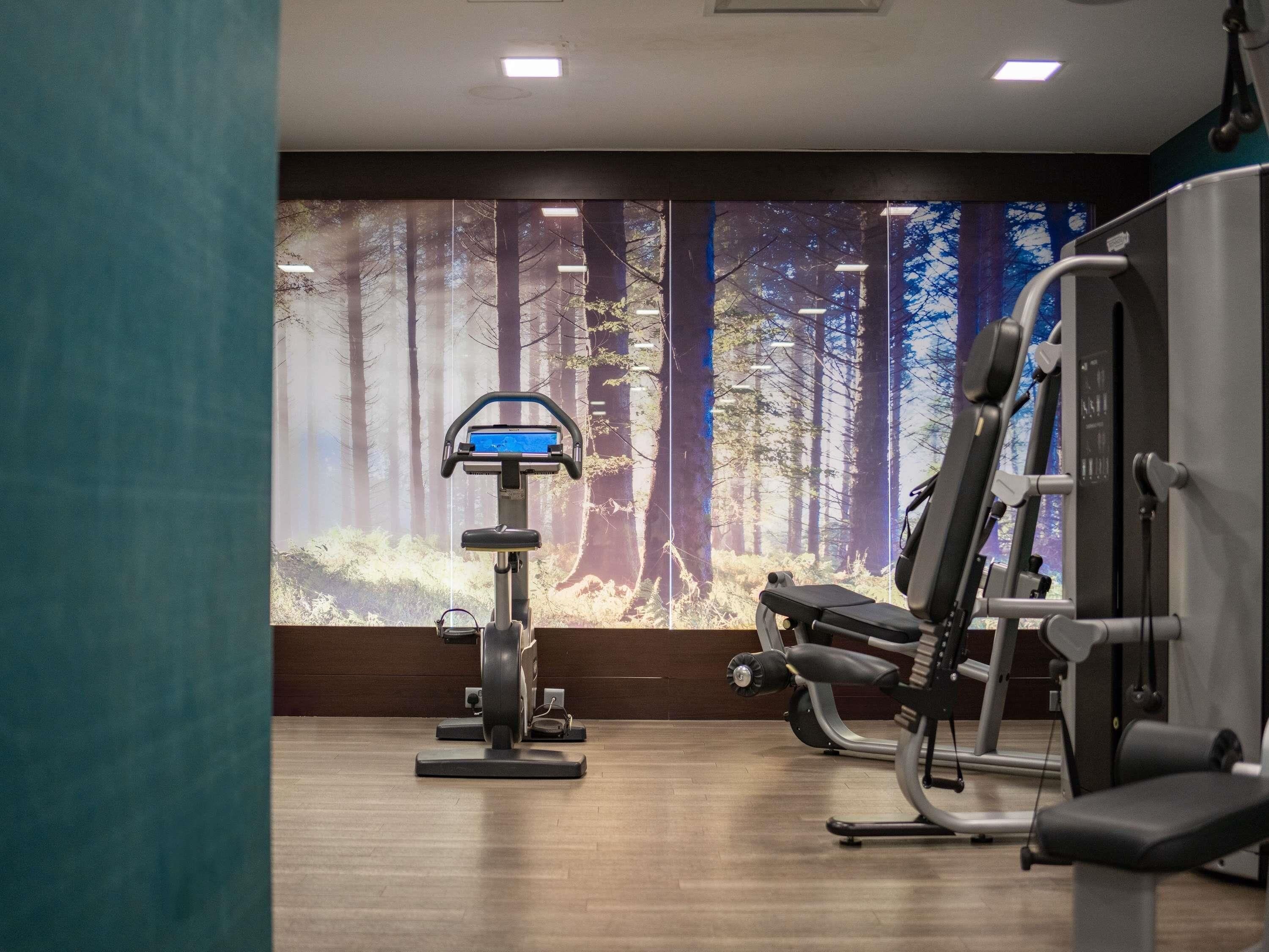 A gym with exercise equipment, a large window with a forest mural, and wooden flooring.