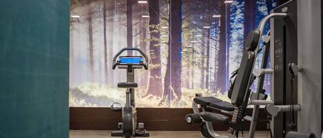 A gym with exercise equipment, a large window with a forest mural, and wooden flooring.