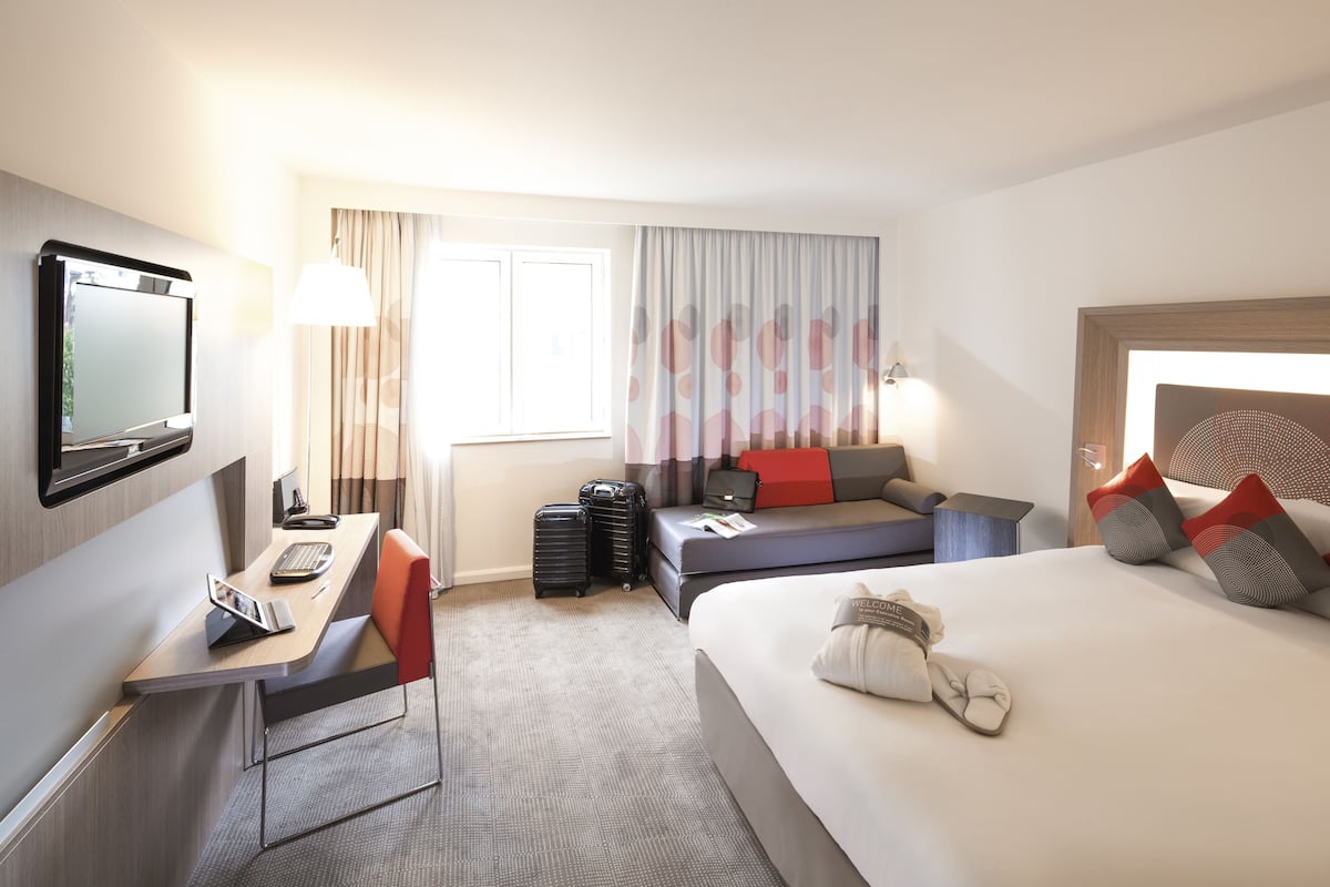 Una habitación de hotel con cama, escritorio y ordenador portátil, silla, televisor, ventana con cortinas y mesita de noche.