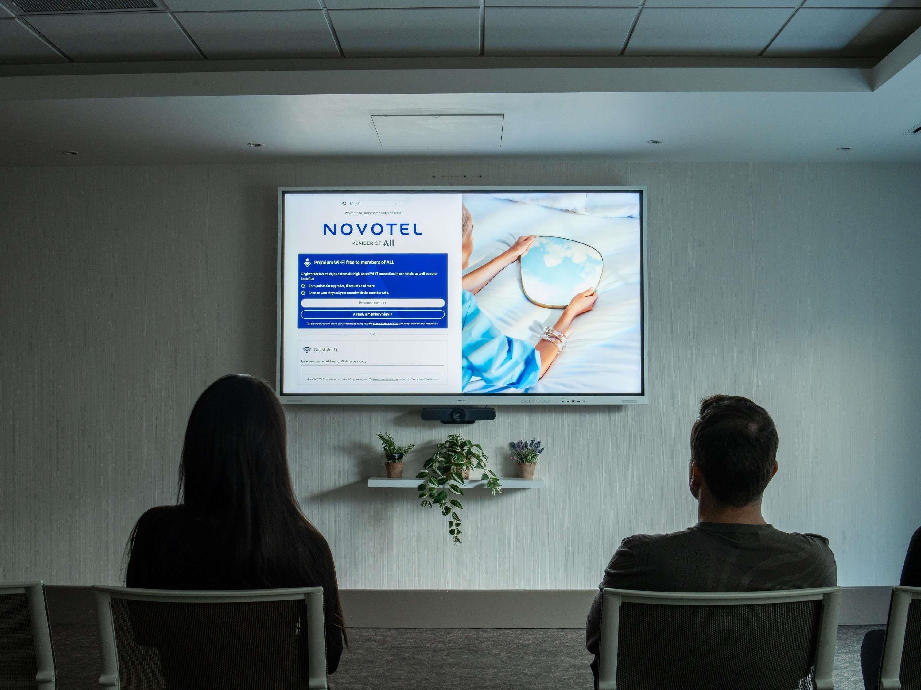 Uno schermo televisivo mostra una pagina di registrazione per il programma fedeltà Novotel, con due persone sedute rivolte verso lo schermo.