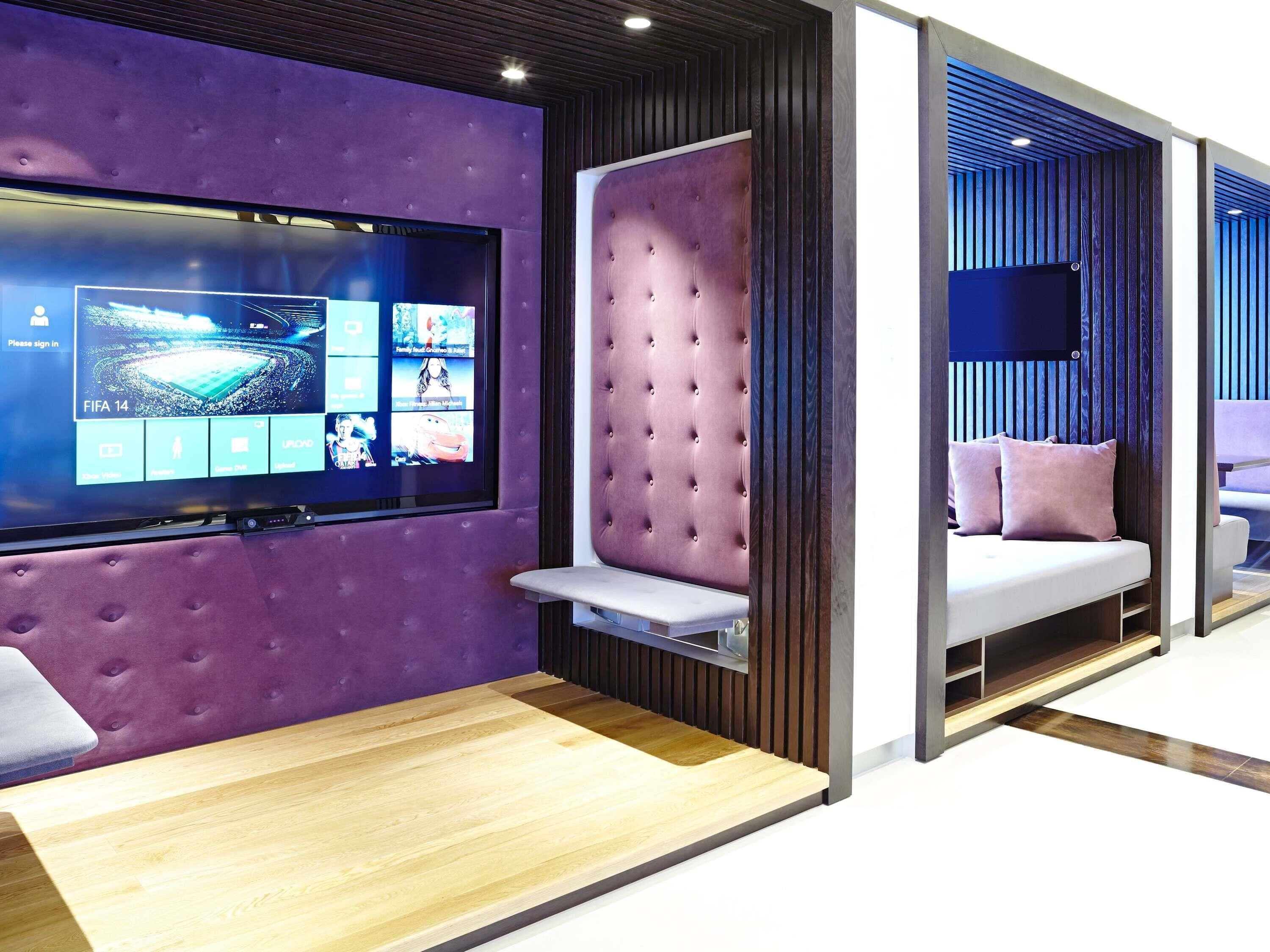 A modern hotel room with a large flat-screen TV, a tufted headboard, a wooden floor, and a seating area with cushions.