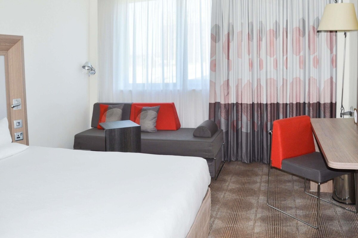 A hotel room with a bed, a sofa with red cushions, a small table, and a chair.