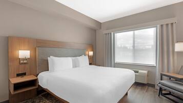 A hotel room with a large bed, a nightstand, a chair, a window with curtains, and a wall-mounted lamp.