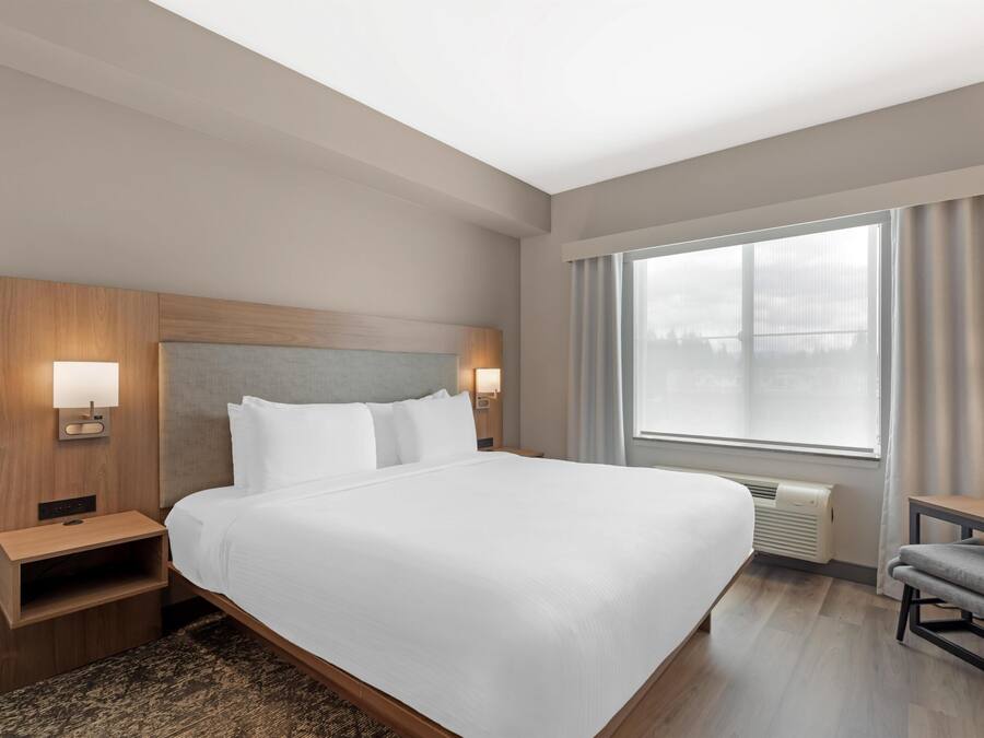 A hotel room with a large bed, a nightstand, a chair, a window with curtains, and a wall-mounted lamp.