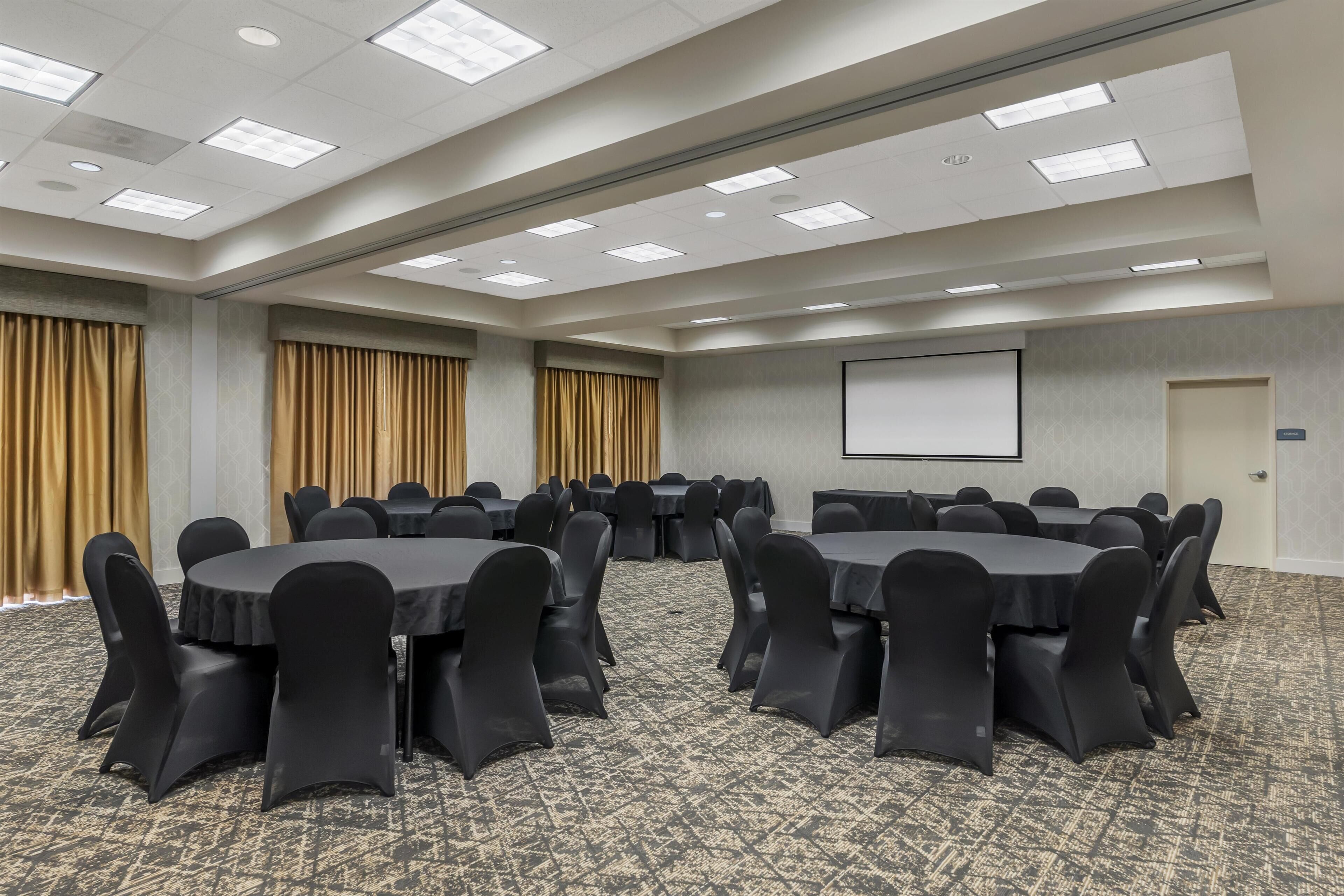 A conference room with round tables, black chairs, a projector screen, and a carpeted floor.