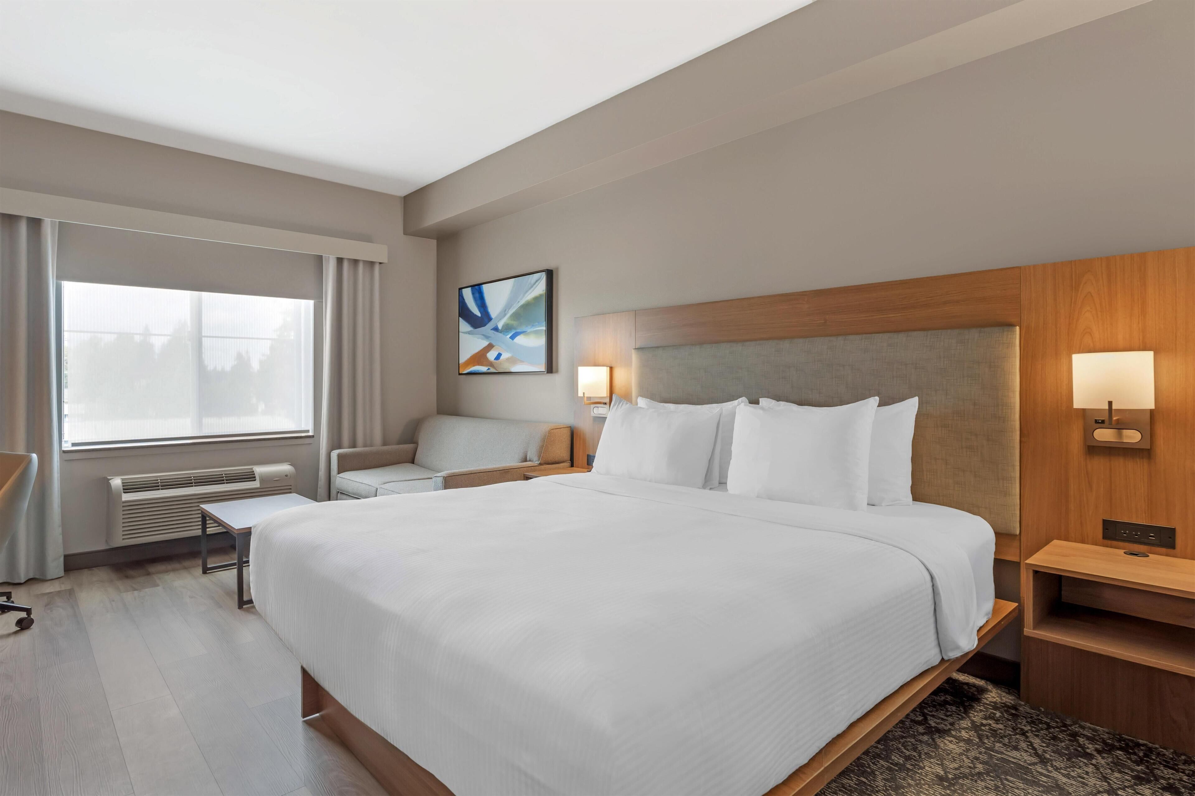 A hotel room with a large bed, a desk, a chair, a window with curtains, a wall-mounted artwork, and a bedside table with a lamp.