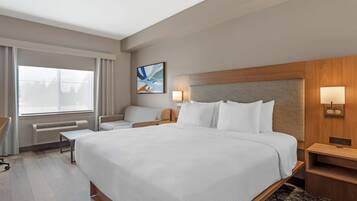A hotel room with a large bed, a desk, a chair, a window with curtains, a wall-mounted artwork, and a bedside table with a lamp.