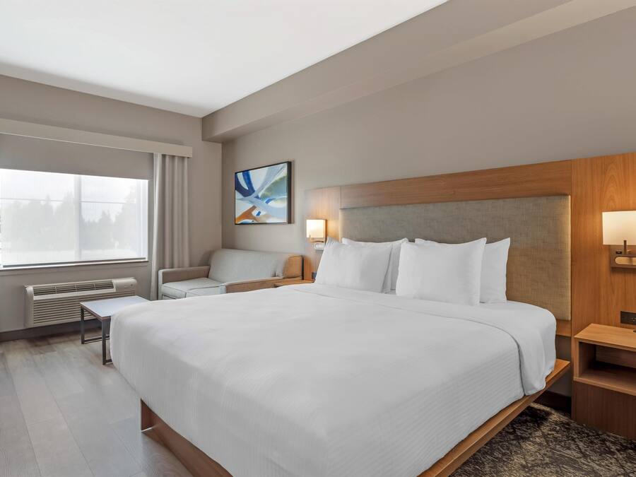A hotel room with a large bed, a desk, a chair, a window with curtains, a wall-mounted artwork, and a bedside table with a lamp.