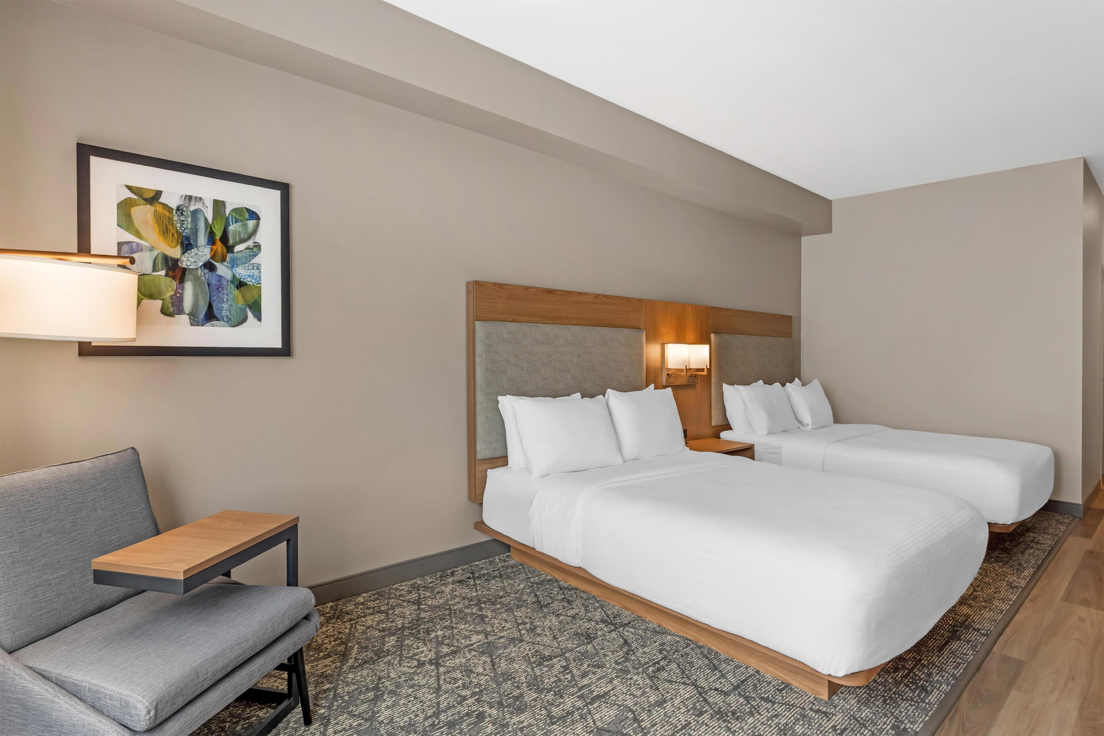 A hotel room with two beds, a desk, a chair, a patterned rug, and a framed artwork on the wall.