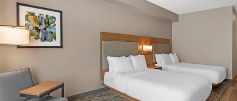 A hotel room with two beds, a desk, a chair, a patterned rug, and a framed artwork on the wall.