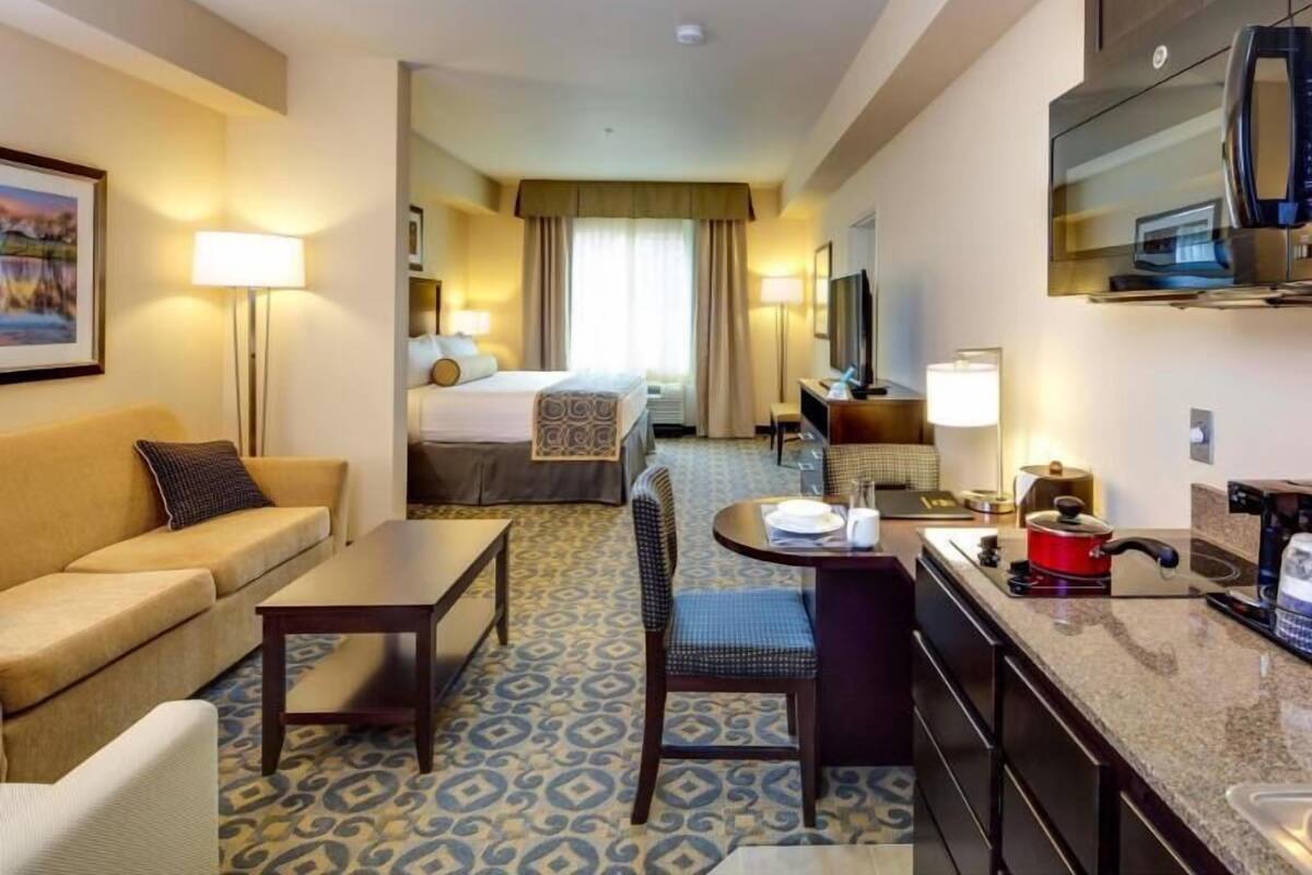 A hotel room with a bed, a sofa, a dining table, a TV, and a kitchen area.