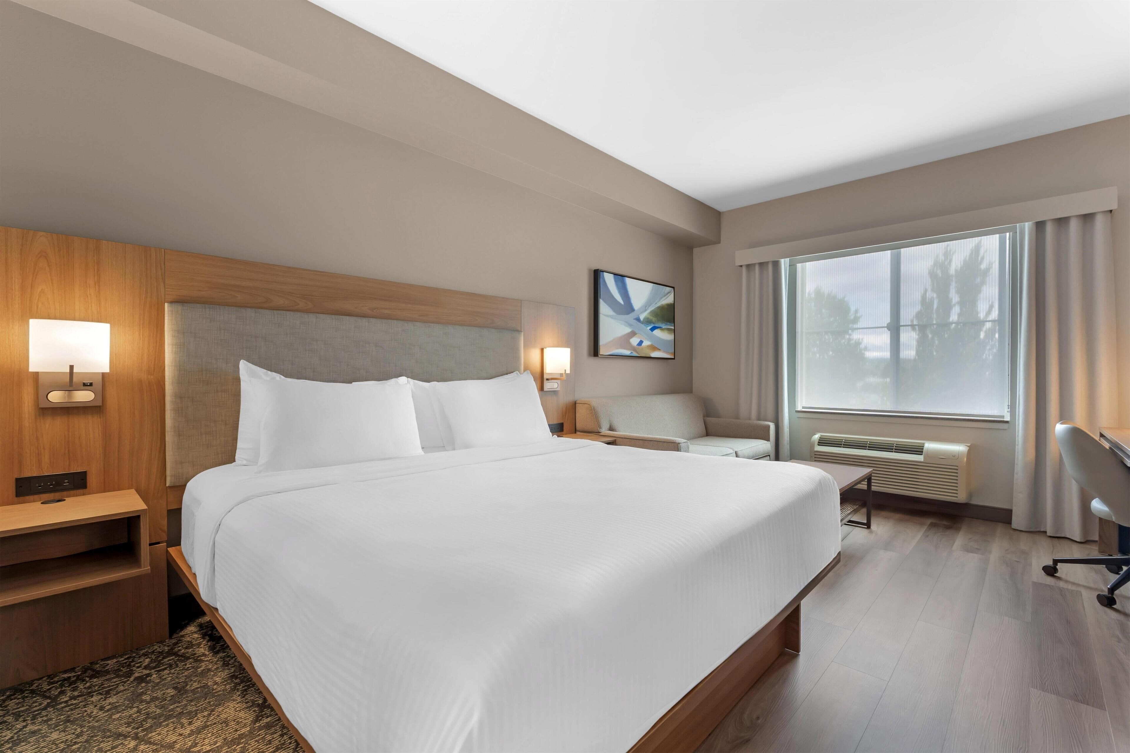 Ein Hotelzimmer mit einem großen Bett, einem Schreibtisch, einem Sessel, einem Fenster mit Vorhängen und einer Wand-Klimaanlage.