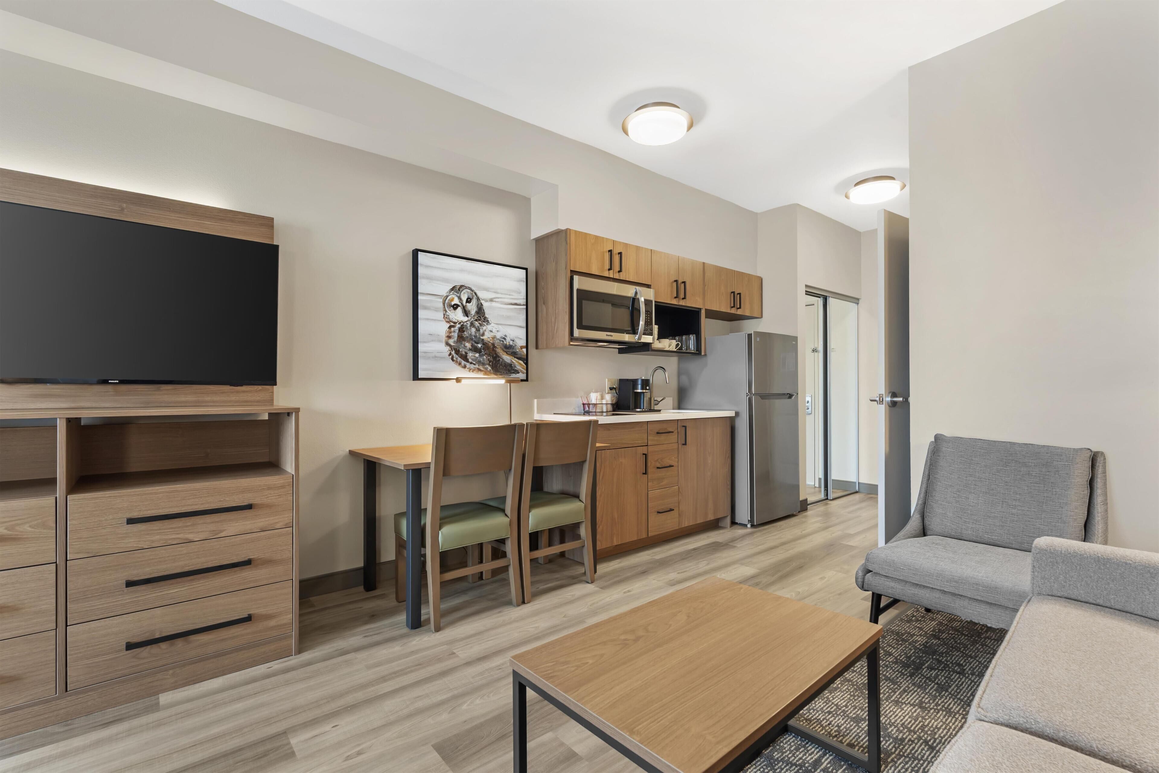 A modern living room with a flat-screen TV, wooden furniture, a dining area with a table and chairs, and a kitchenette with a microwave and refrigerator.