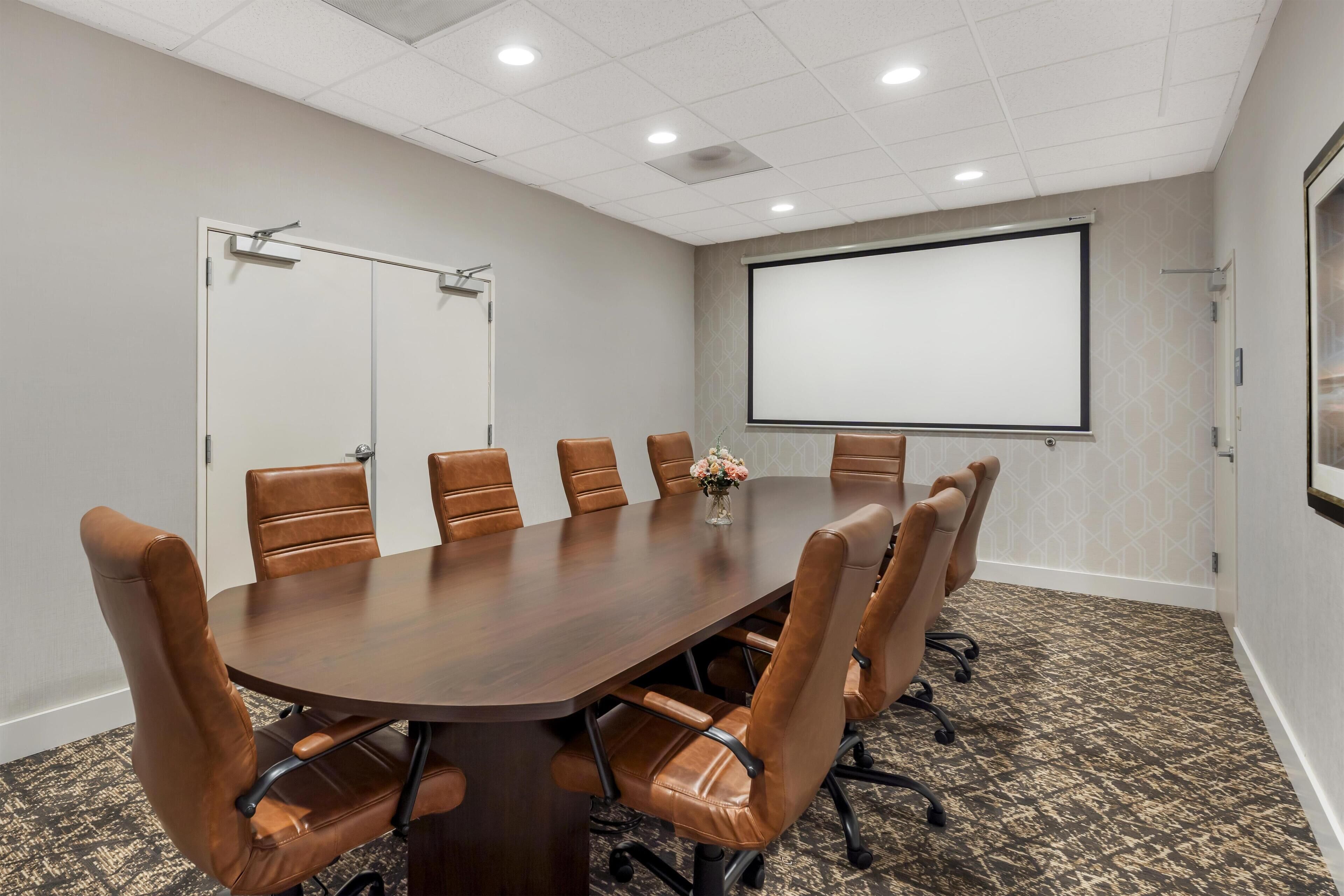 A conference room with a large wooden table, brown leather chairs, a projector screen, and a carpeted floor.