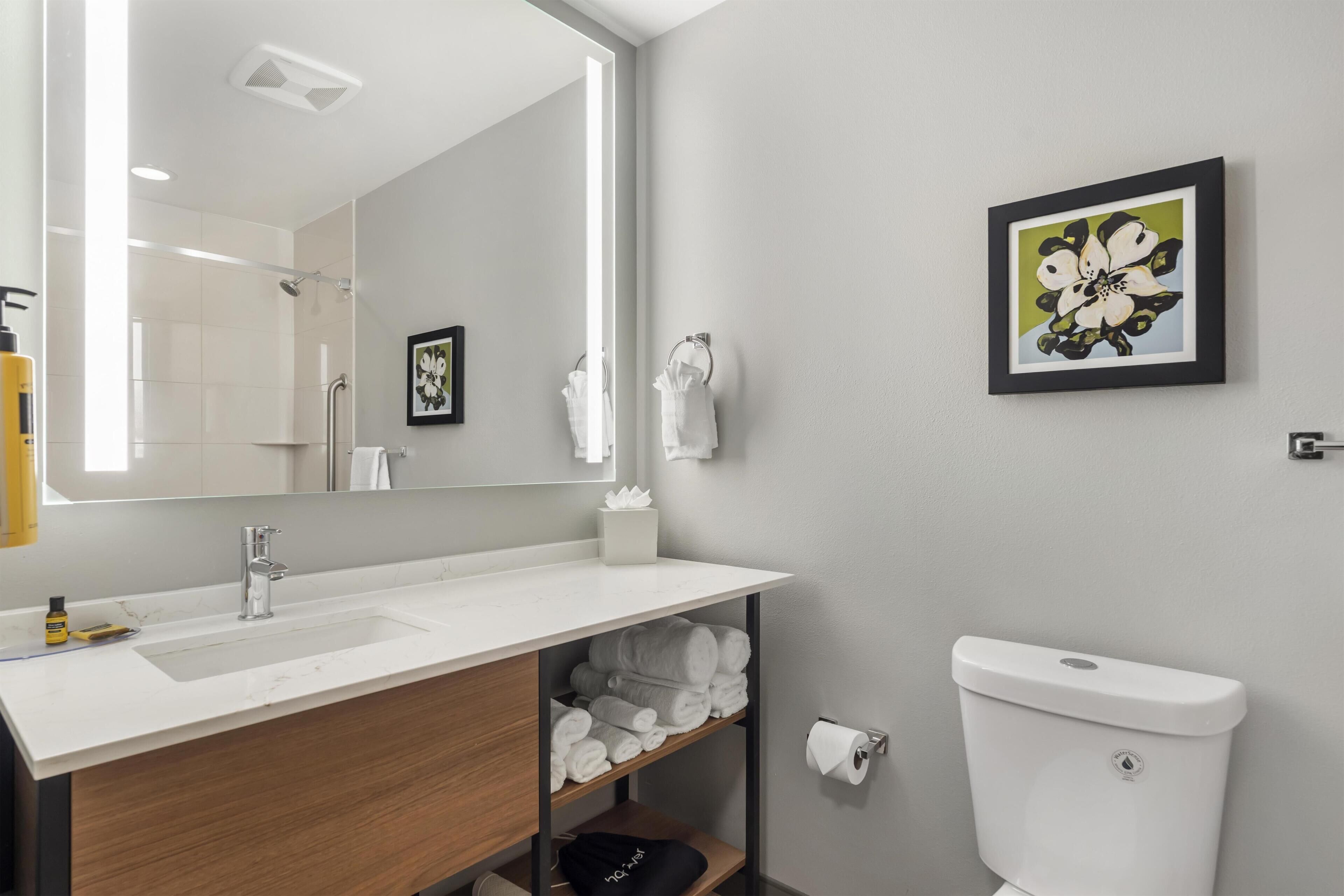 A modern bathroom with a large mirror, a sink, a toilet, and a shelf with towels.