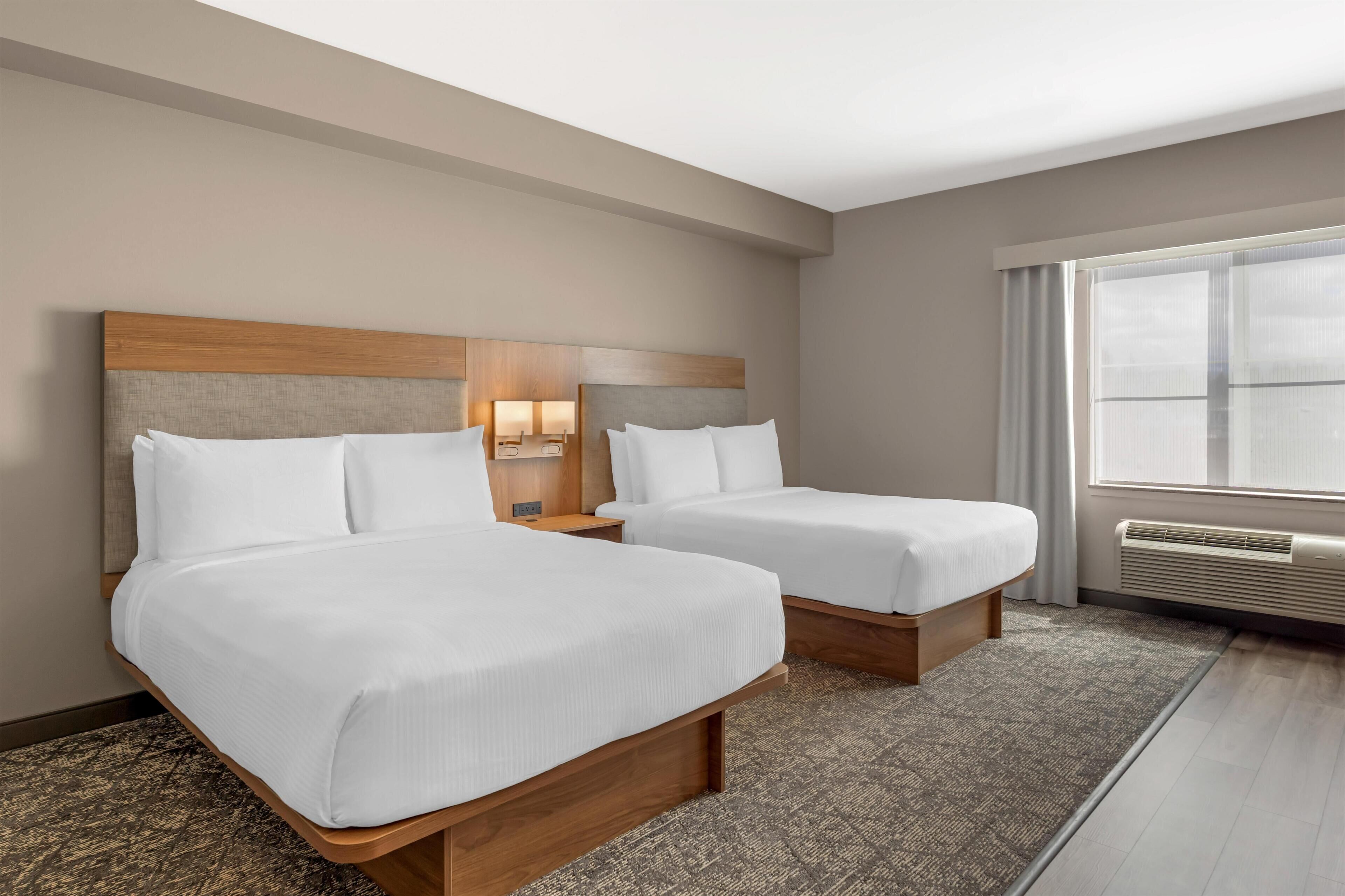 A hotel room with two beds, a nightstand, a lamp, and a large window with curtains.