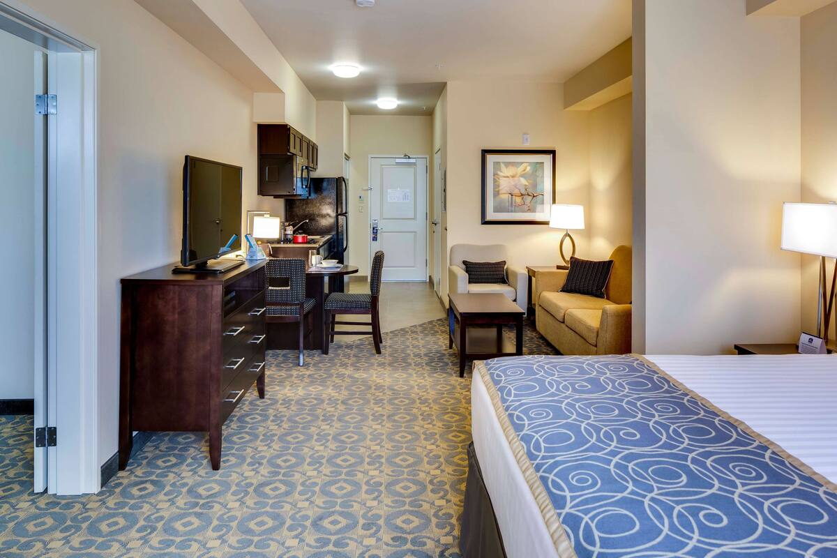 A hotel room with a bed, a desk, a chair, a sofa, a TV, a kitchen area, and a framed picture on the wall.