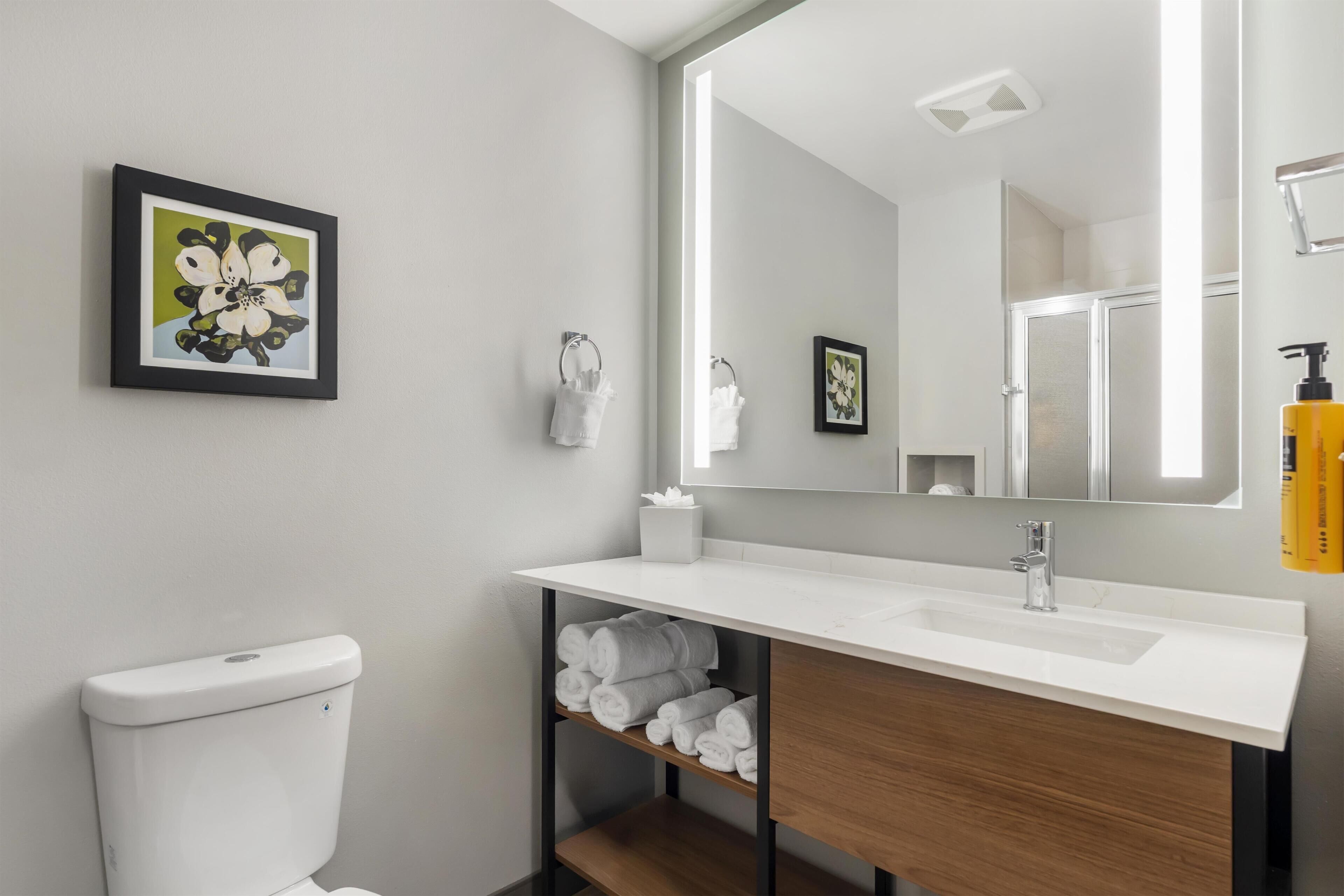 Un baño moderno con inodoro blanco, lavabo con tocador de madera, espejo y repisa con toallas enrolladas.
