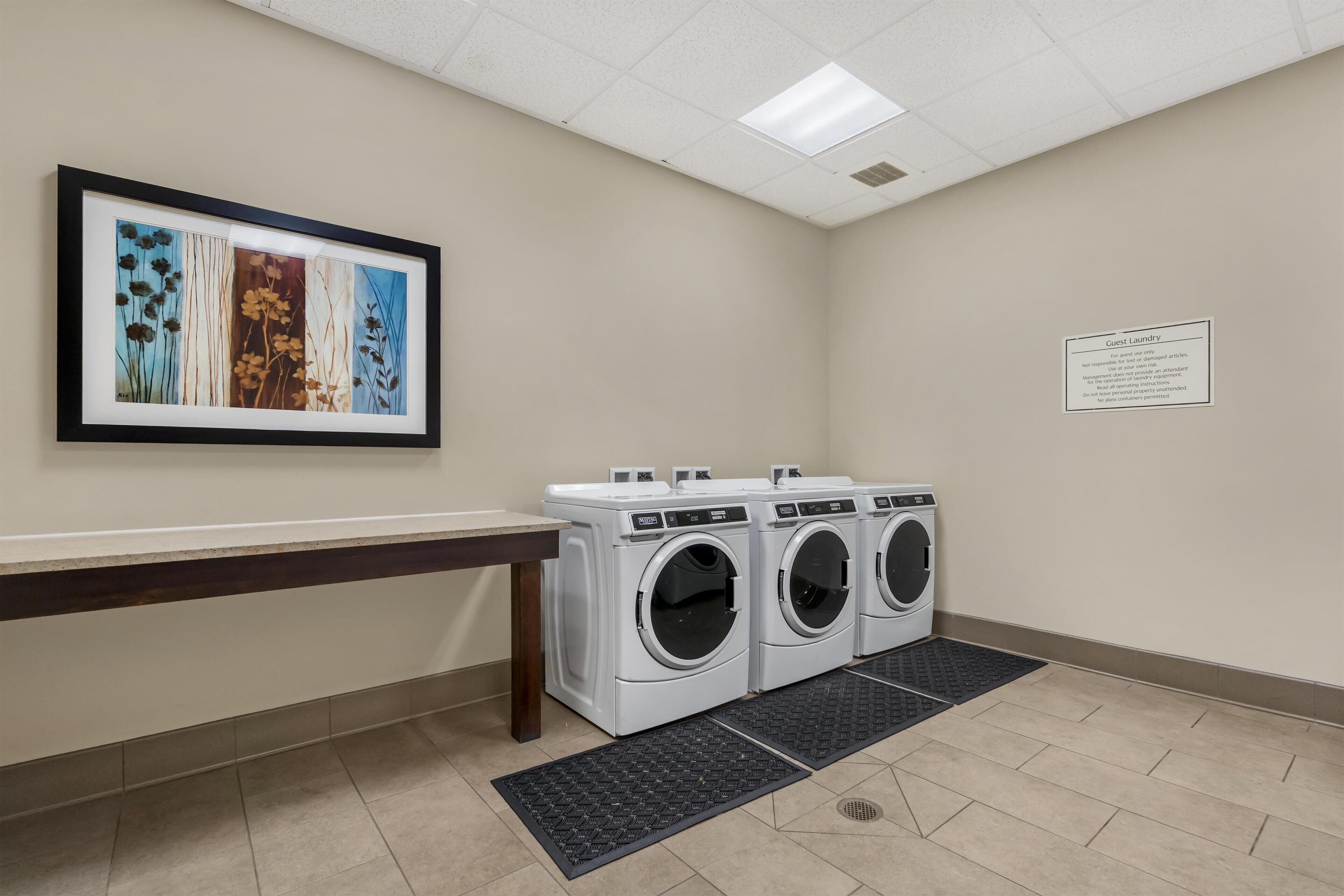 Une buanderie équipée de trois machines à laver et de trois sèche-linge superposés, d’une œuvre d’art encadrée accrochée au mur et d’un panneau indiquant les services de blanchisserie mis à la disposition des clients.