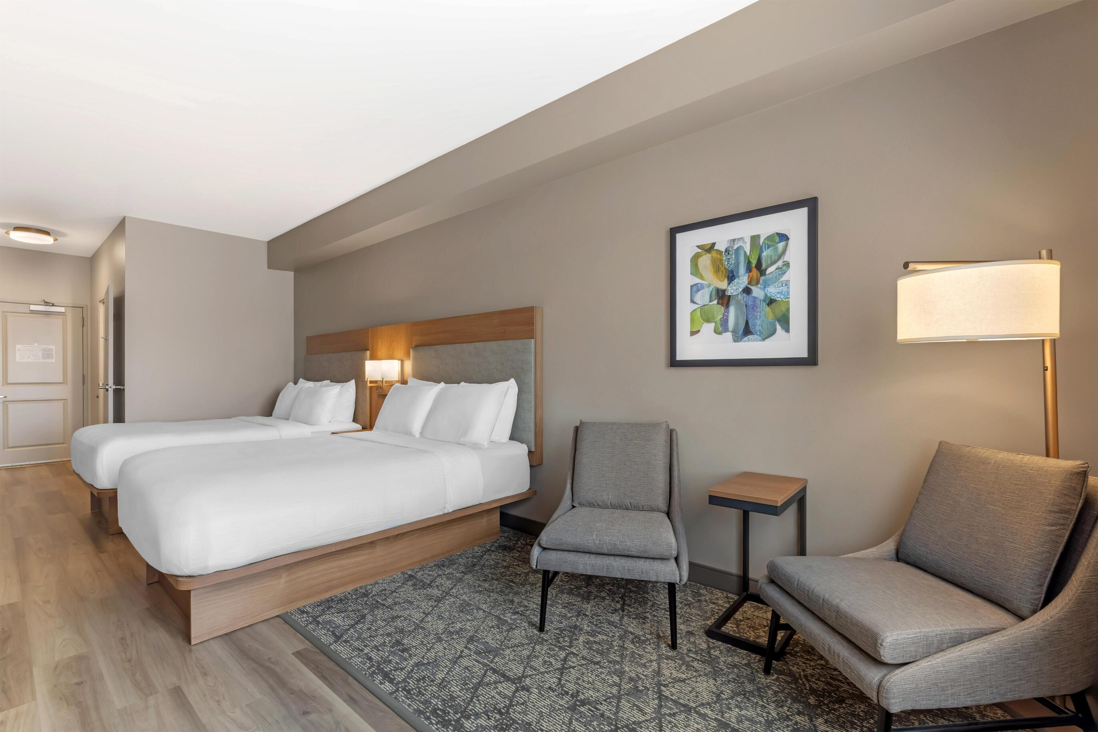 A hotel room with two beds, a chair, a small table, a lamp, and a framed picture on the wall.