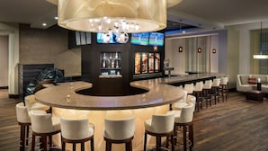 A bar area with a round counter, multiple bar stools, a large chandelier, and a television screen displaying sports.