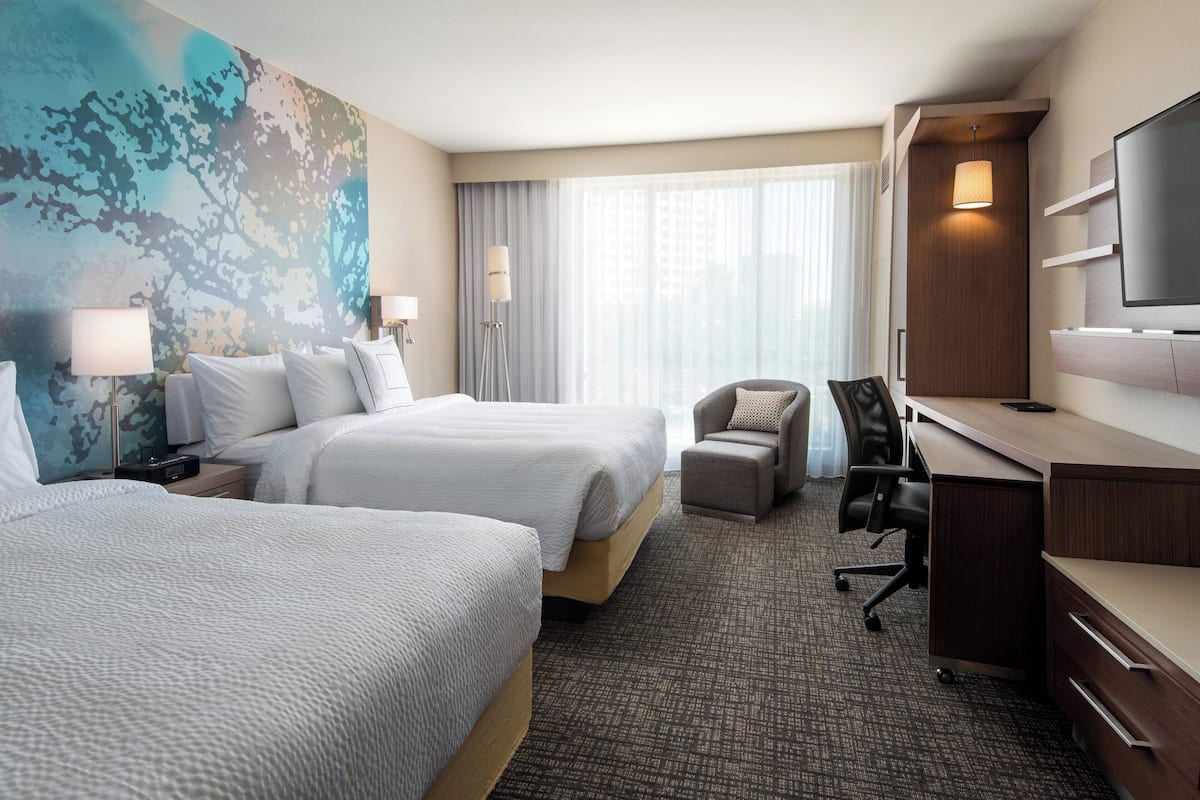 A hotel room with two beds, a desk, a chair, a TV, and a large window with curtains.