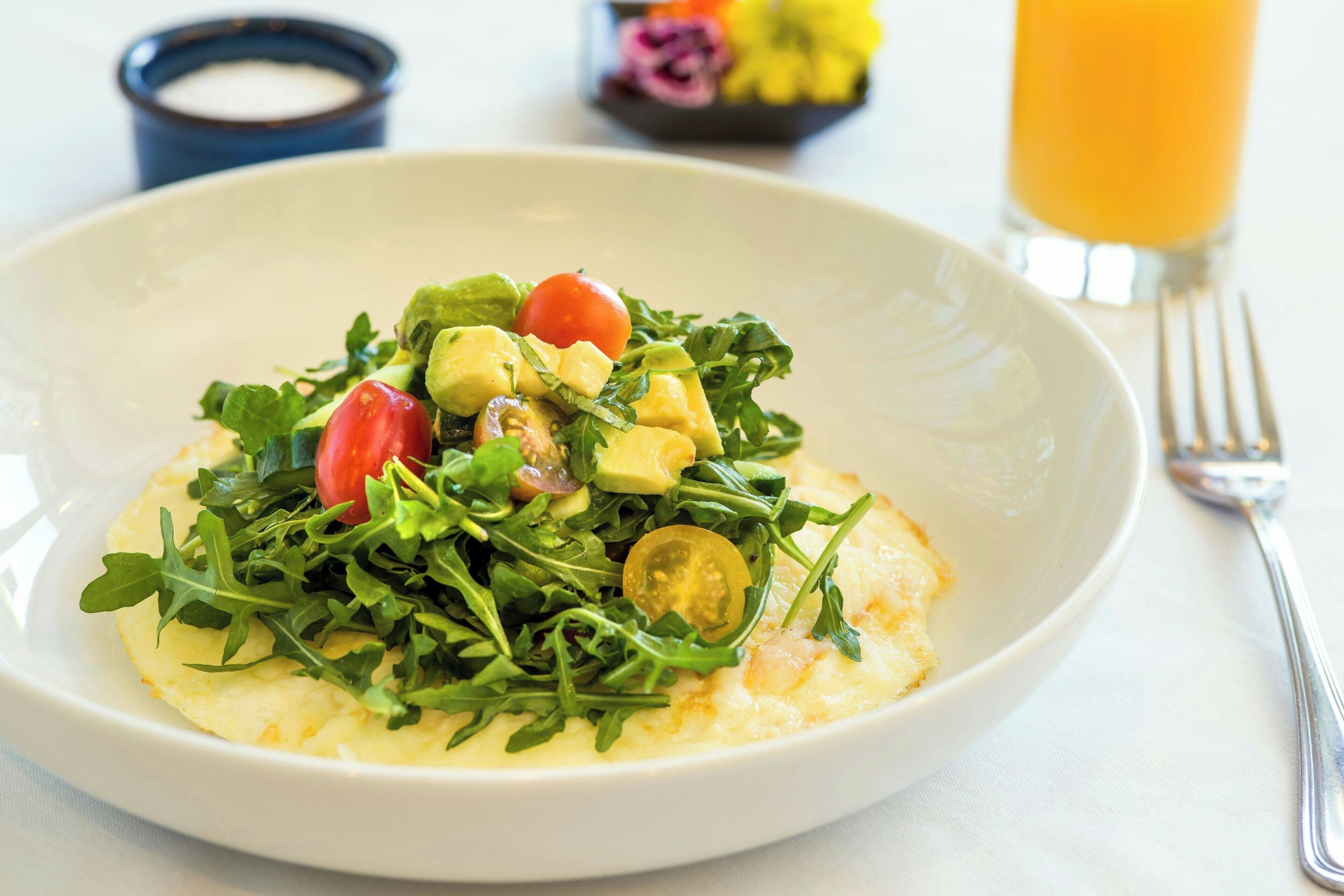 A plate of food with greens, tomatoes, and avocado on top of a creamy base, possibly scrambled eggs, with a glass of orange juice and a fork on the side.